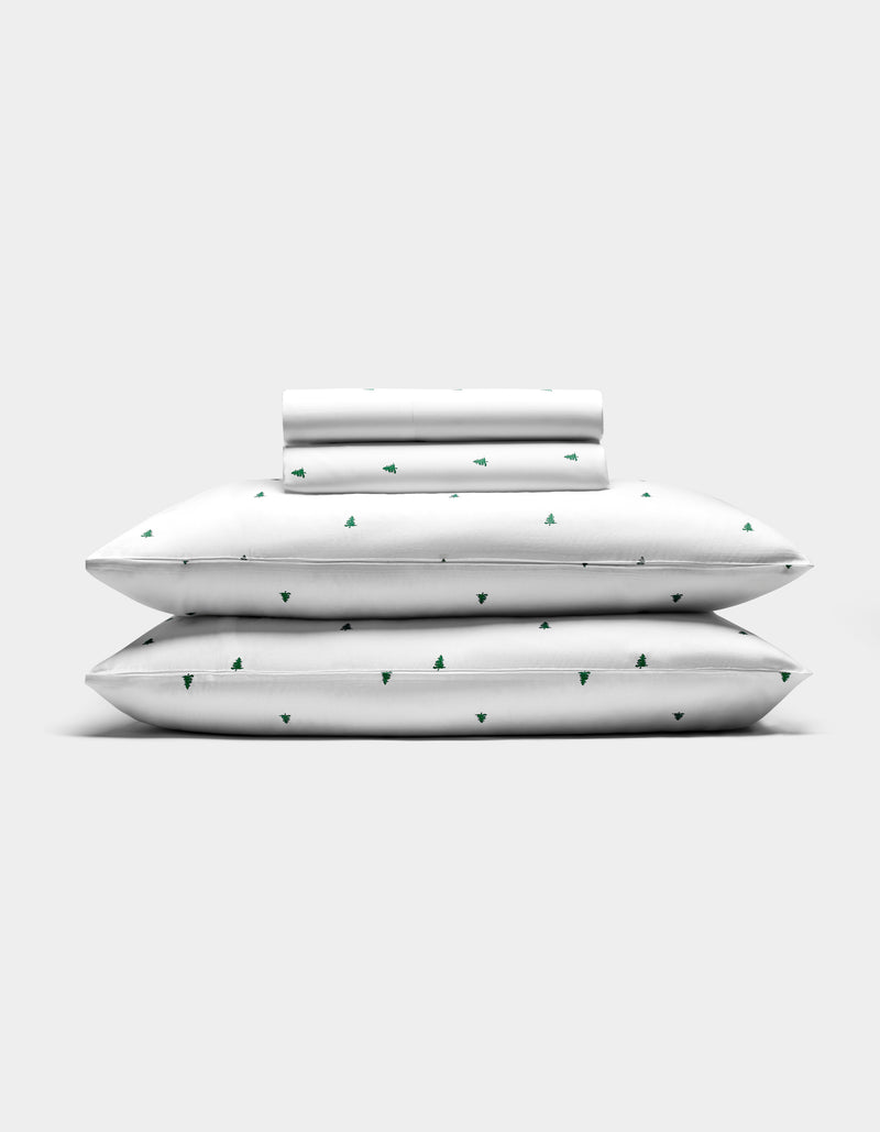 The Cozy Earth Bamboo Sheet Set features two pillows, a folded sheet, and a rolled pillowcase—all white with small green tree patterns—arranged against a plain light gray background.