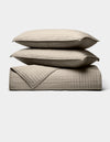 The Cozy Earth Bamboo Jersey Quilt Bundle, shown against a white background, features two beige quilted pillows stacked on a matching blanket with a simple square-stitched design.|Color:Driftwood