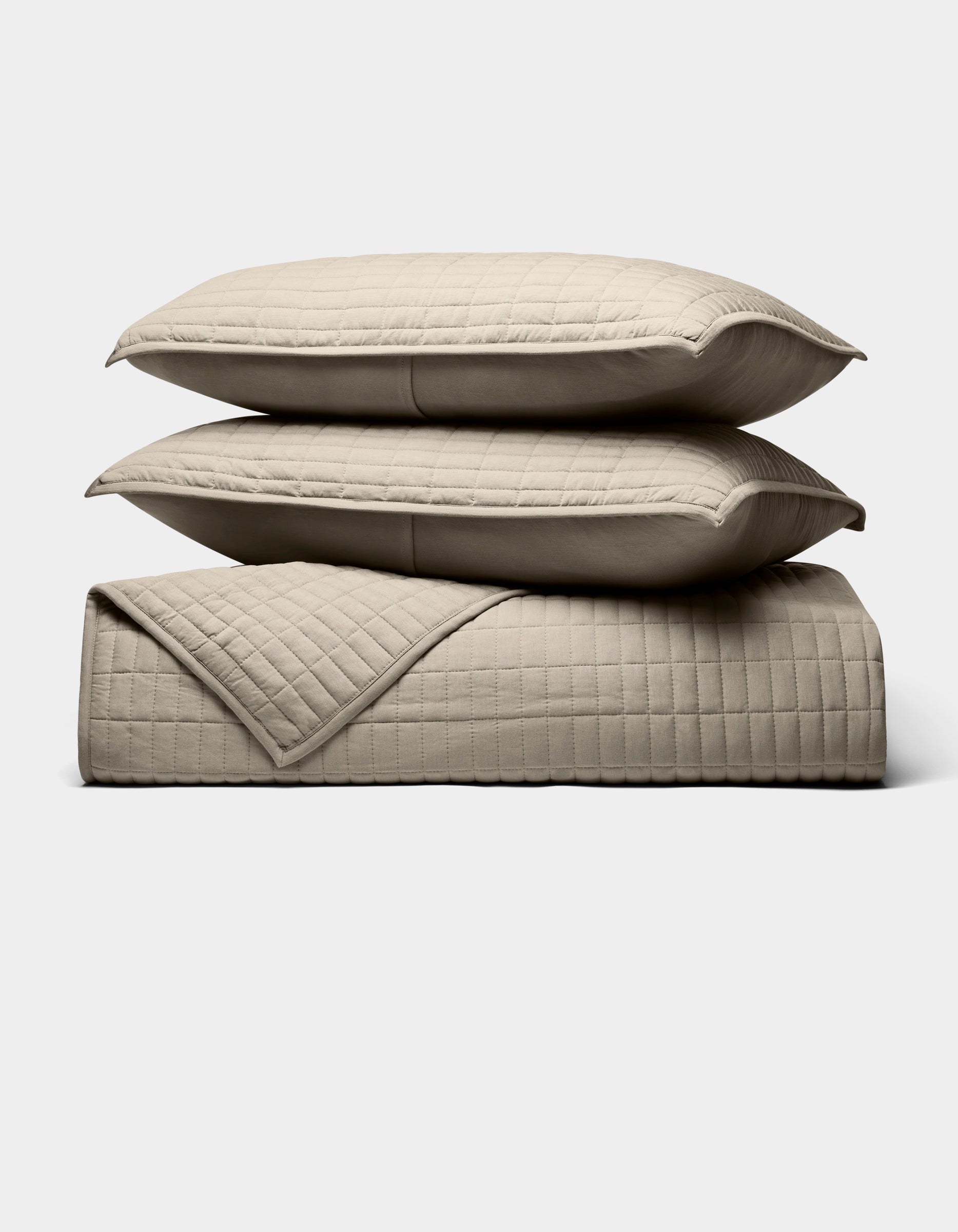 The Cozy Earth Bamboo Jersey Quilt Bundle, shown against a white background, features two beige quilted pillows stacked on a matching blanket with a simple square-stitched design.|Color:Driftwood