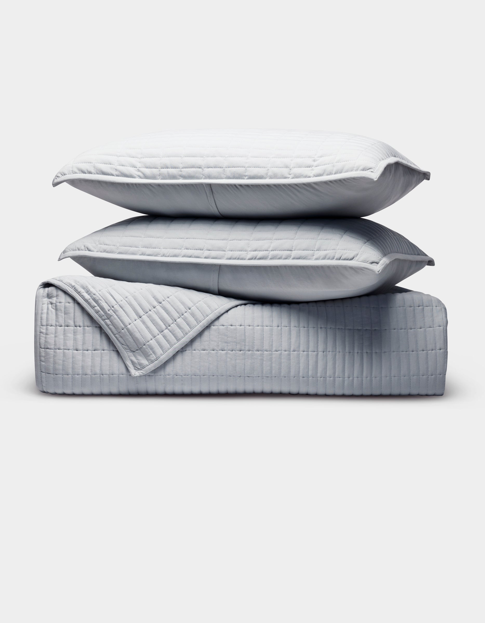 The Bamboo Jersey Quilt Bundle by Cozy Earth features a neatly folded light gray quilted comforter with matching pillows stacked on top, all displayed against a clean white background.|Color:Shore