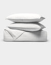 The Cozy Earth Bamboo Jersey Quilt Bundle features two white pillows stacked on a folded white quilted blanket, all with a subtle grid pattern, set against a plain light gray background.|Color:White