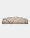 The Cozy Earth Bamboo Jersey Quilt, featuring a beige, quilted grid pattern, is neatly folded on a white background. A turned-down corner highlights its soft, luxurious texture.|Color:Driftwood