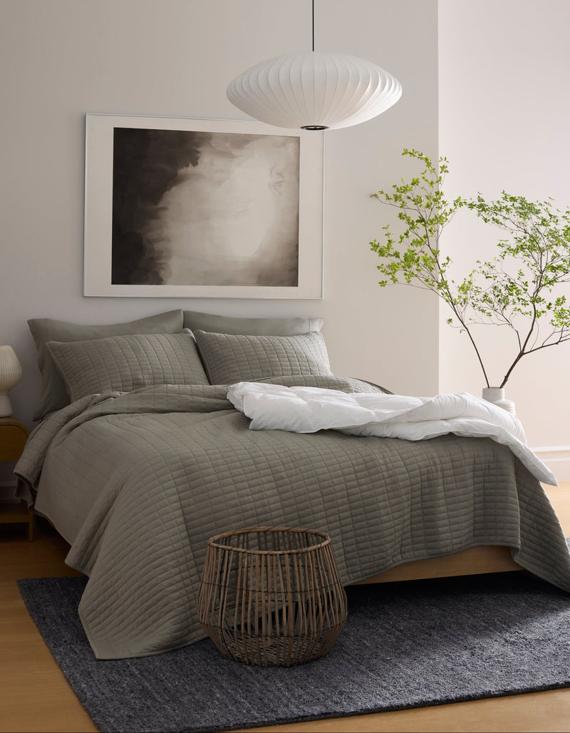 A modern bedroom features a neatly made bed with the Cozy Earth Bamboo Jersey Quilt, a large abstract artwork above the headboard, a paper lantern ceiling light, a green leafy plant, a woven basket, and a dark rug on light wood flooring.