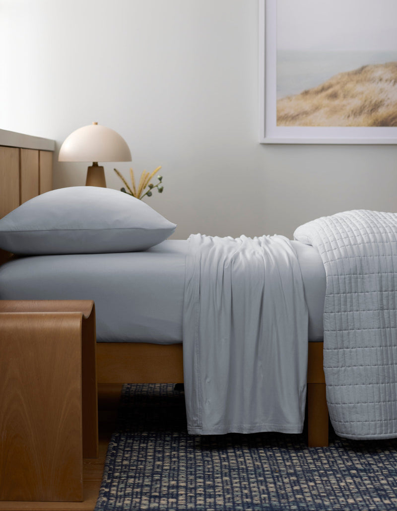 A neatly made bed with the Cozy Earth Bamboo Jersey Sheet Set in light gray sits in a minimalist room. Above the bed hangs a framed landscape photo, while a wooden bench and a lamp complete the look over a patterned blue rug.