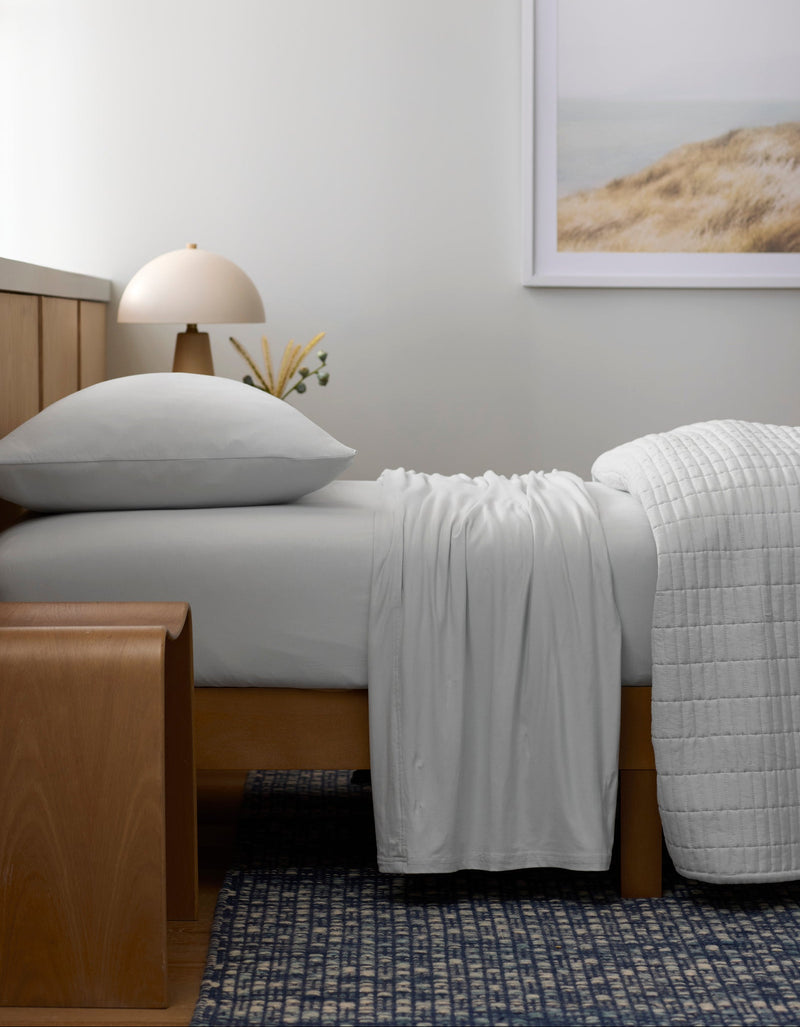 A bed made with Cozy Earth’s Bamboo Jersey Sheet Set rests against a wooden headboard. A wooden bench stands at the foot, while a lamp, small vase, and framed landscape photo accentuate the minimal, serene bedroom.