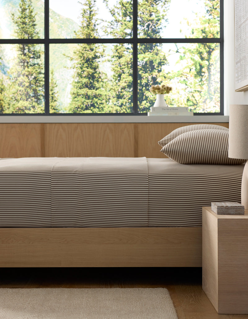 A neatly made bed with Cozy Earth’s Bamboo Fitted Sheet in beige and white stripes sits by a wooden nightstand in a modern bedroom, with large windows framing tall green trees and mountains outside.