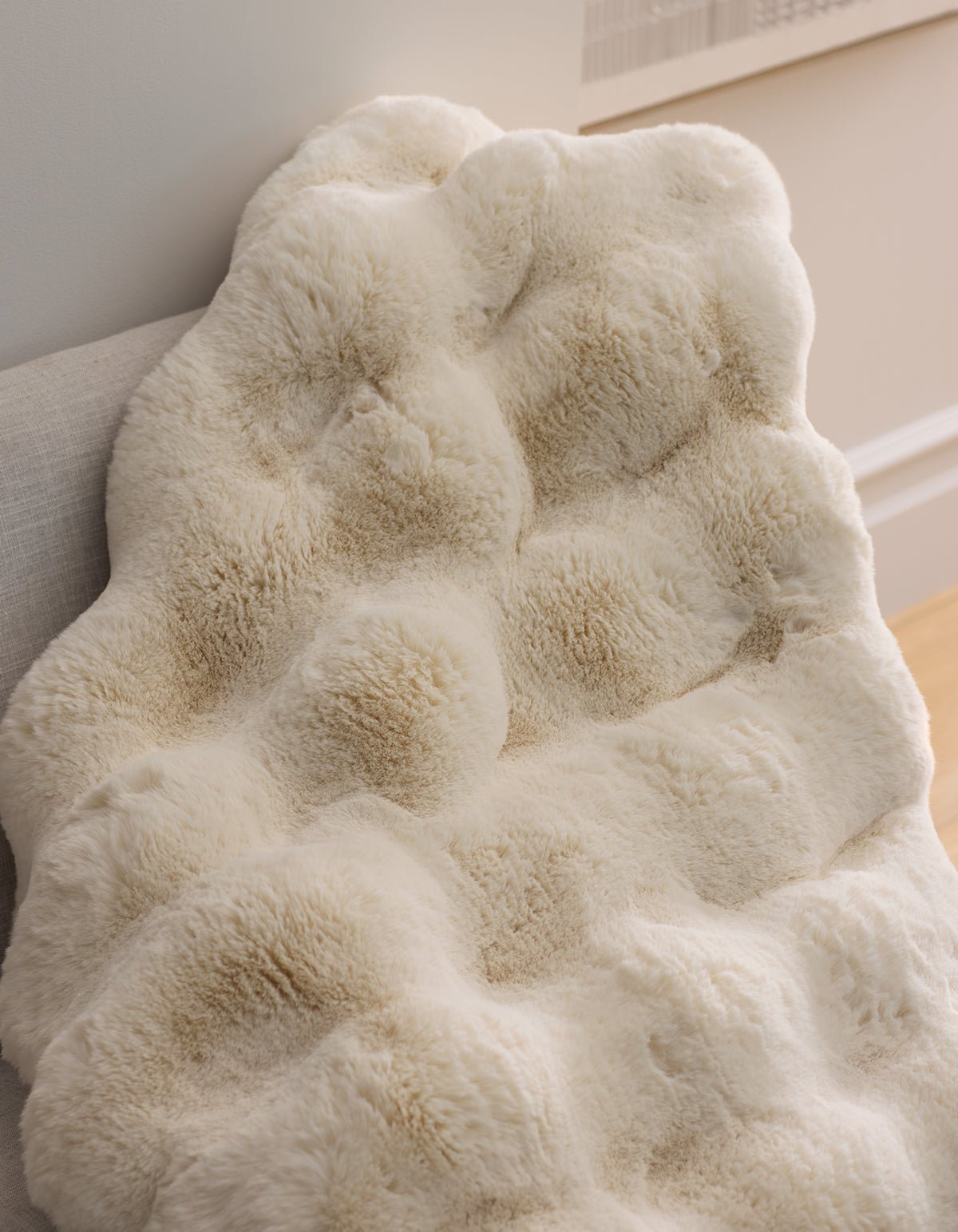 The Cozy Earth Double Bubble Cuddle Blanket, featuring a cream-colored faux fur with a textured, tufted design, is draped over a light chair in a stylish, comfortable room.|Color:Creme