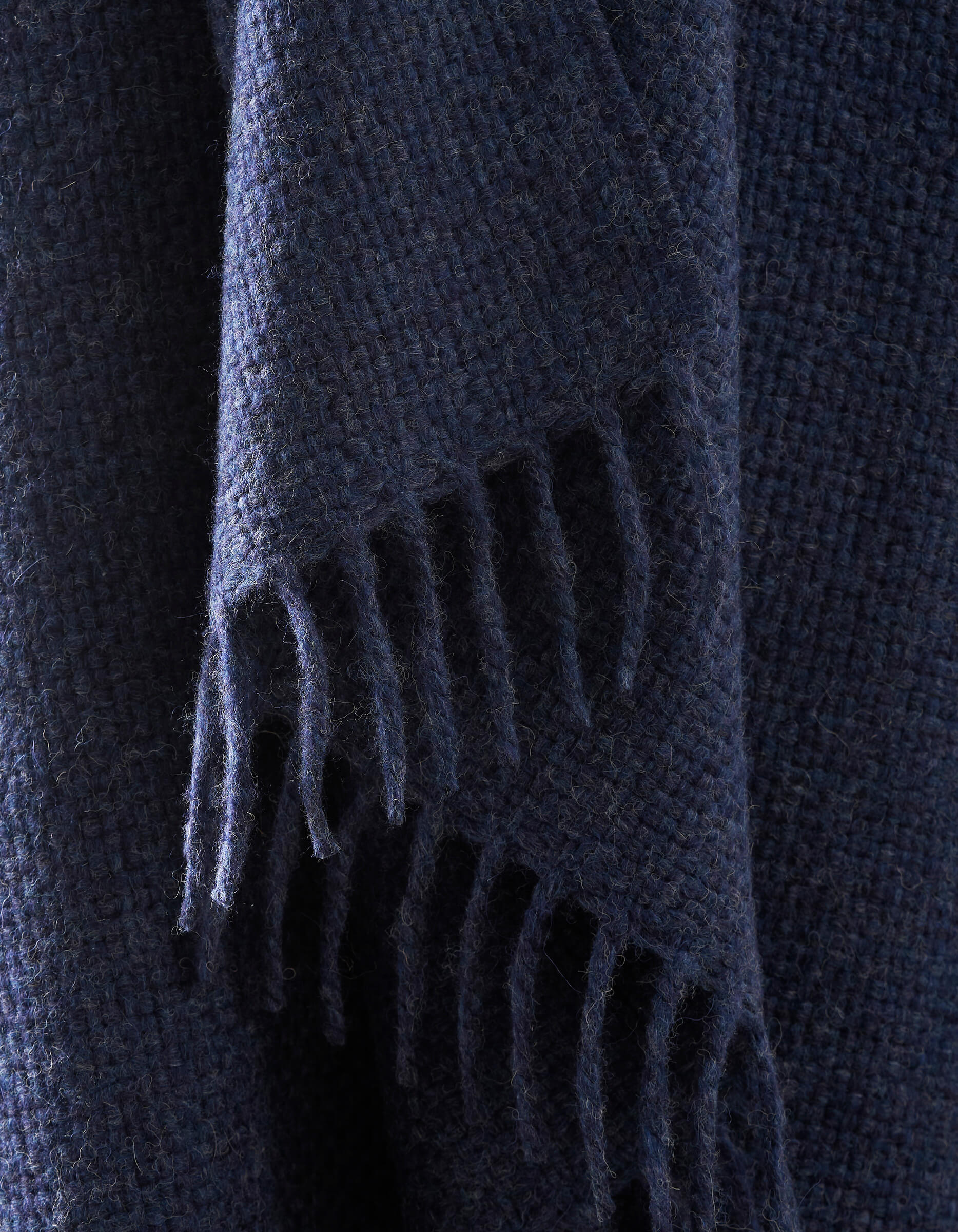 A close-up of the Lucia Throw by Cozy Earth shows its dark blue, textured woven fabric with fringed edges hanging down.