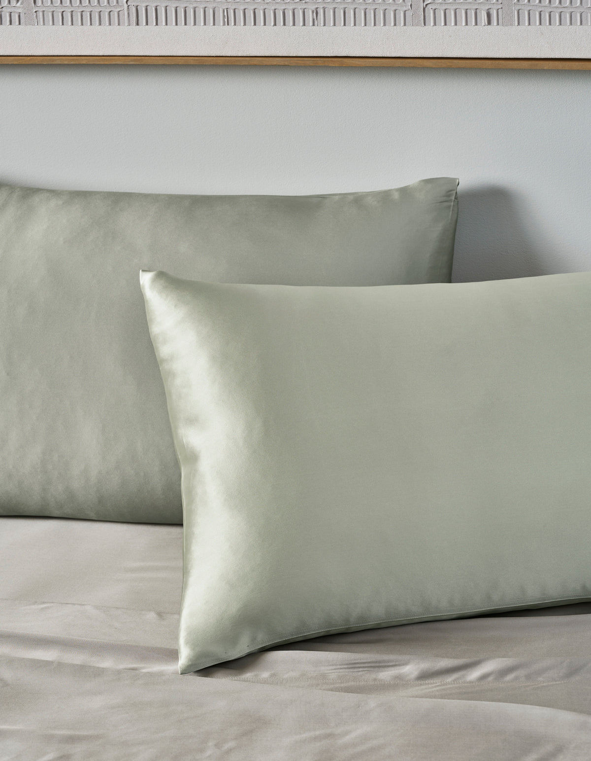 Two Cozy Earth Silk Pillowcases in pale green rest on a matching sheet, set against a light gray wall with part of a wooden frame visible above.|Color:Laurel