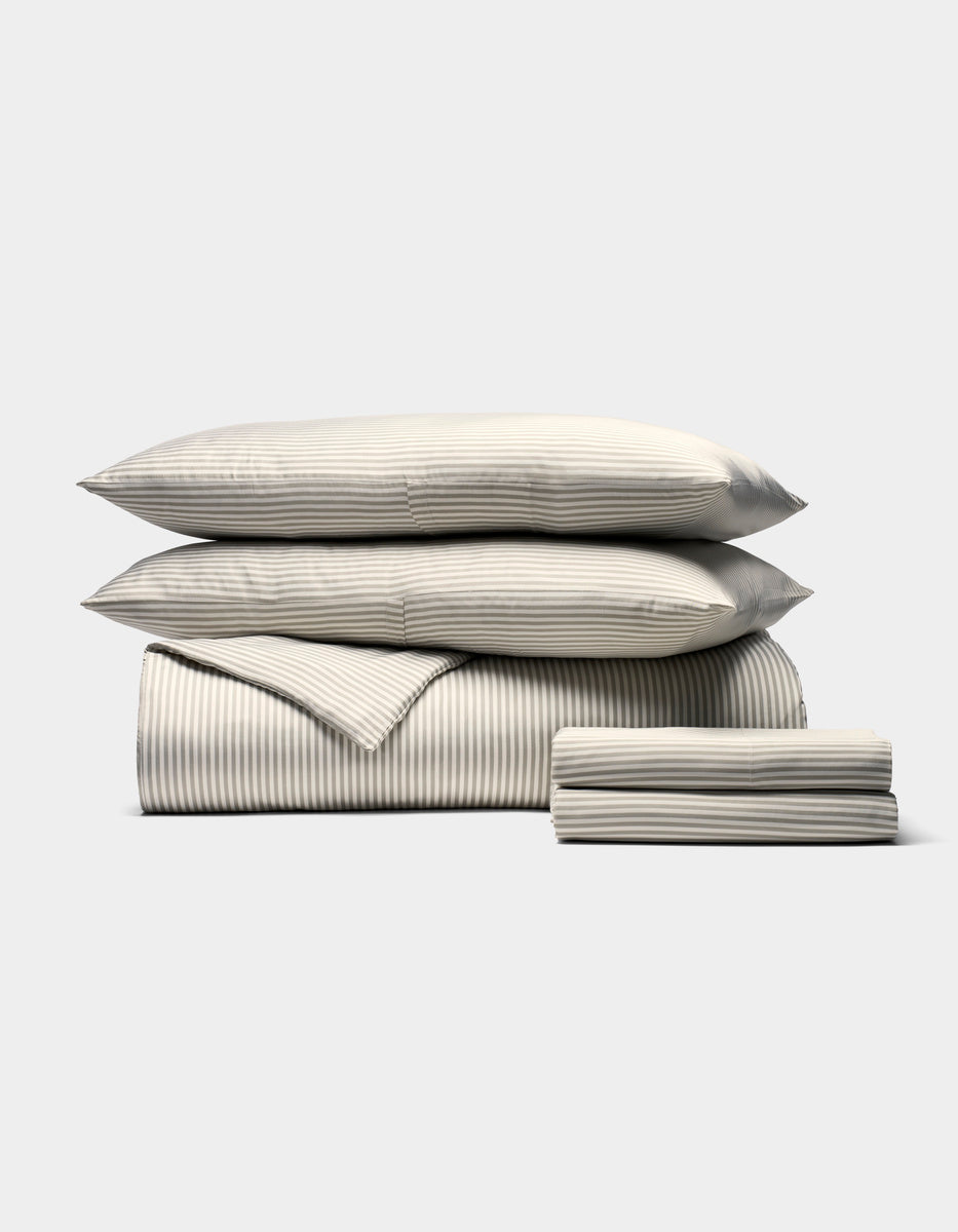 The Bamboo Bedding Deluxe Bundle by Cozy Earth is shown neatly stacked with two pillows, a folded comforter, and sheets in a subtle light gray and white pinstripe pattern against a plain background.