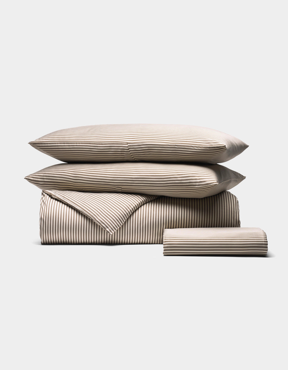 The Cozy Earth Bamboo Bedding Essential Bundle features beige and white striped bedding—two pillows, a folded duvet, and a folded sheet—neatly displayed against a plain light gray background.|Color:Classic Stripe in Ebony