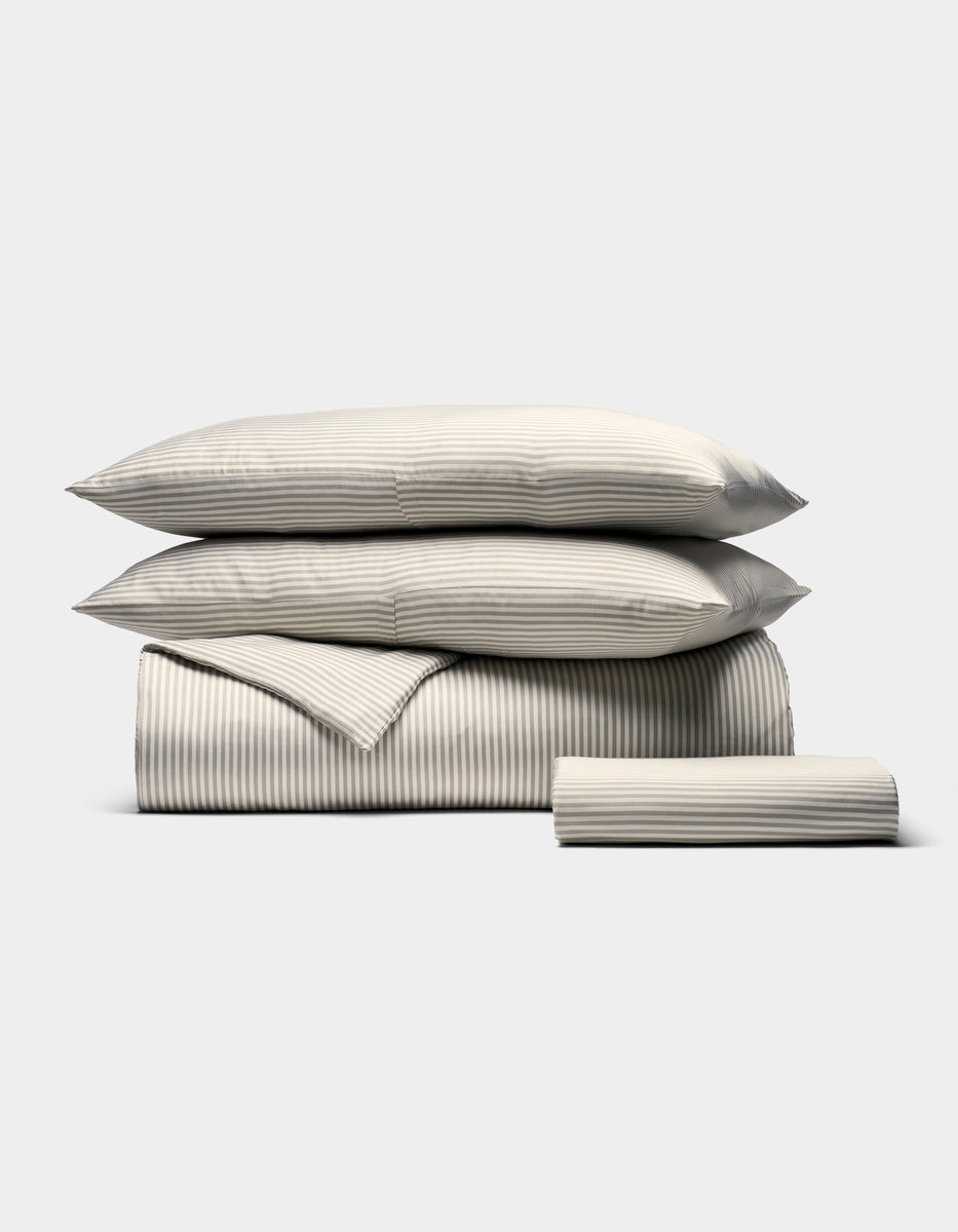 The Bamboo Bedding Essential Bundle by Cozy Earth features a neatly stacked set of beige and white striped bedding—two pillows, a folded duvet, and a folded sheet—displayed on a plain light background.|Color:Classic Stripe in Laurel
