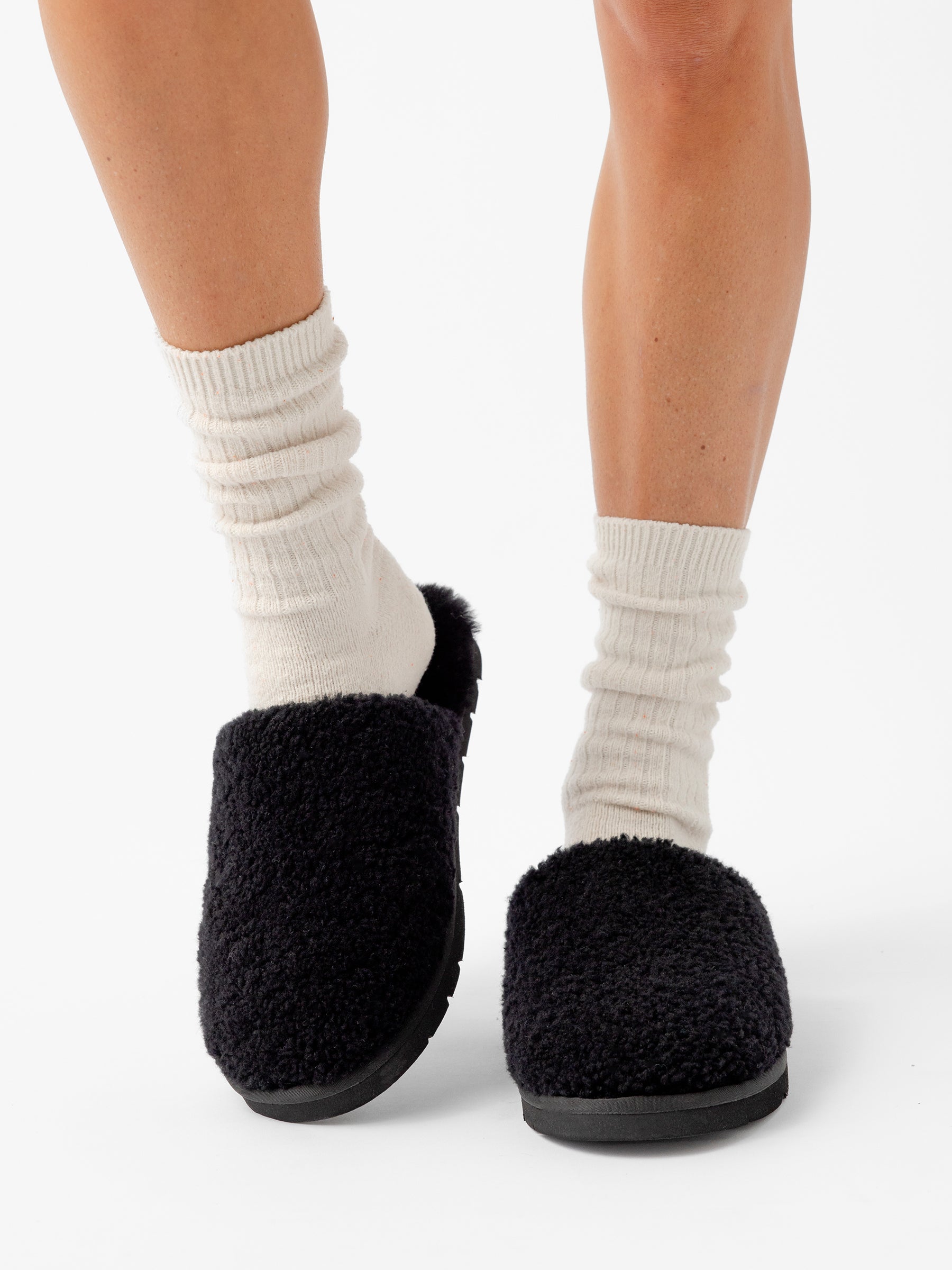 A person wearing white, ribbed socks and Cozy Earth's Puffy Sheep Slippers is standing on a white background. Only their lower legs and feet are visible. The slippers have a cozy, textured appearance that contrasts with the light-colored socks. |Color:Black Sheep