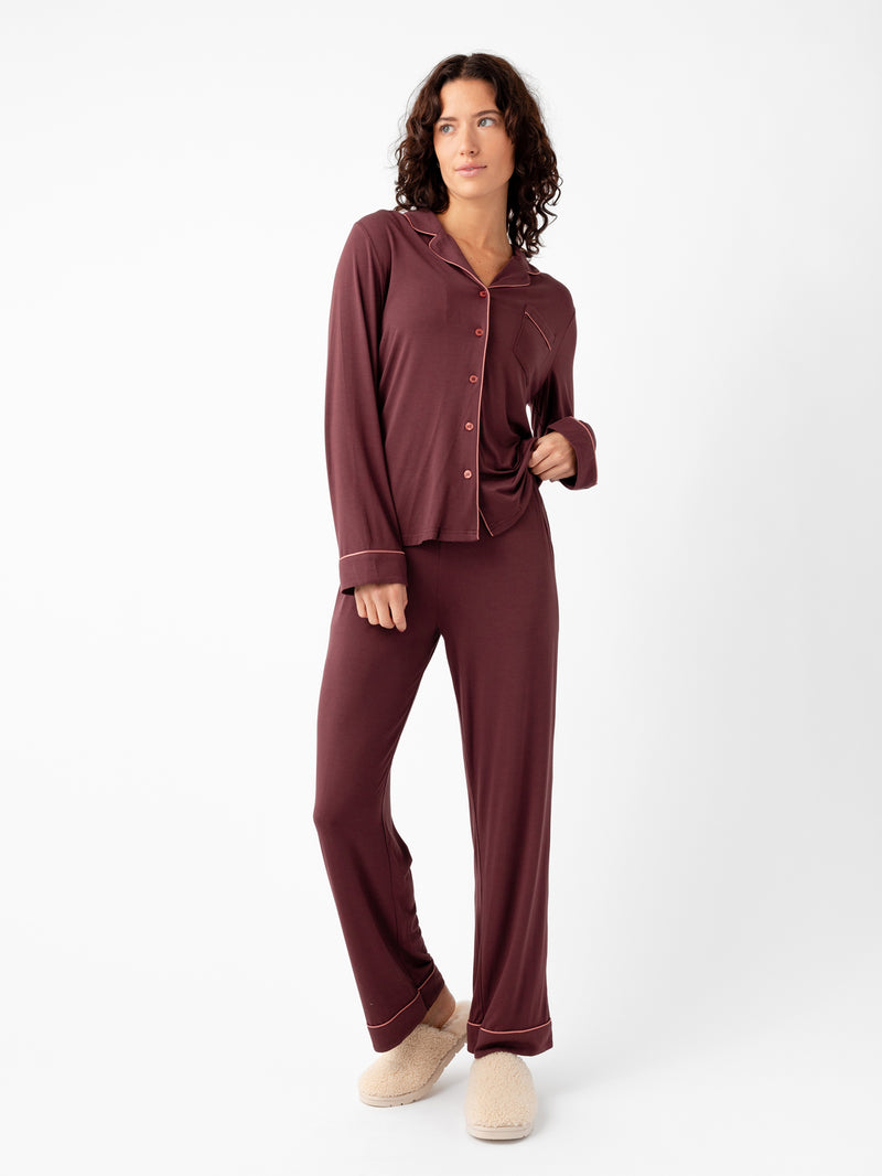 A person with curly hair wears the Cozy Earth's Women's Bamboo Stretch-Knit Long Sleeve Pajama Set in maroon, featuring a button-up top and matching pants, paired with beige slippers. The backdrop is plain white.