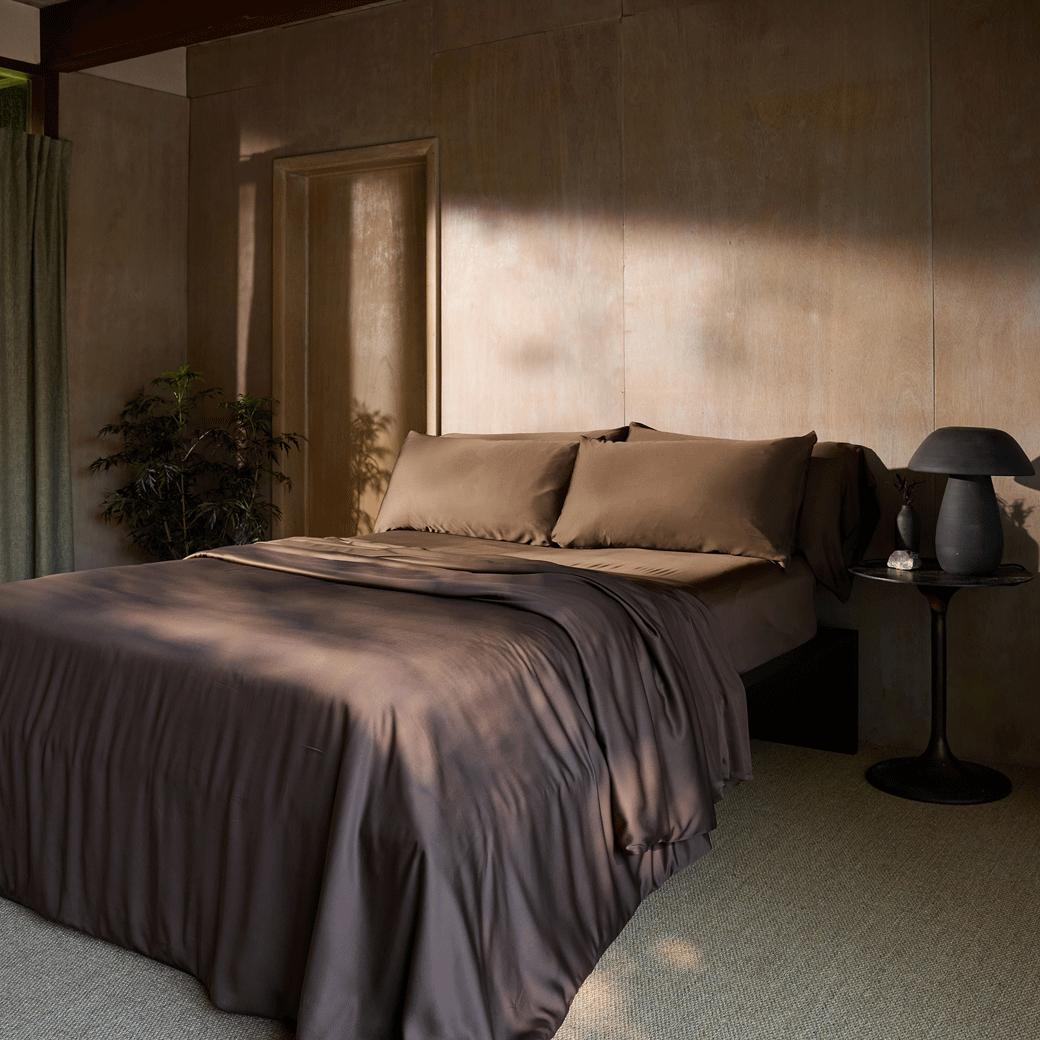 A bed dressed in Cozy Earth’s Bamboo Fitted Sheet - Last Chance sits in a modern, minimalist bedroom with light wood-paneled walls, a round black side table, lamp, and potted plant. Sunlight streams in through the window.