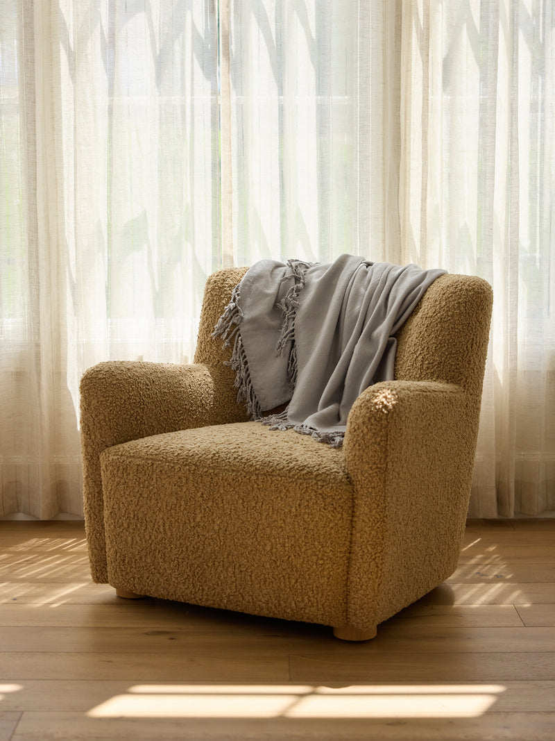 A cozy beige armchair with textured fabric is placed in front of a sheer, sunlit curtain. A light gray Bamboo Tassel Throw by Cozy Earth is draped casually over the backrest, and sunlight casts soft shadows on the wooden floor.