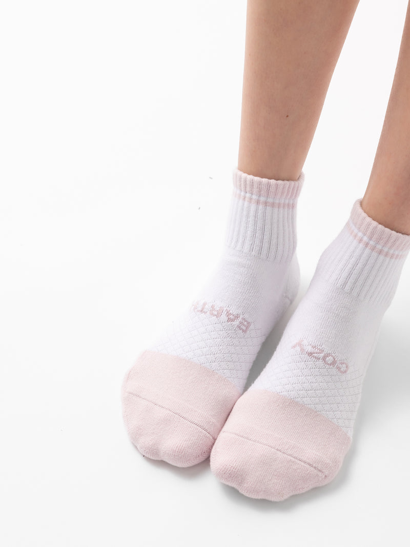 A person's lower legs are wearing a pair of Cozy Earth's Essential Quarter Sock from the 2-Pack collection. The white socks feature light pink toes and heels, ribbed cuffs, and a small, subtle pink logo near the top of each foot. The background is plain white.