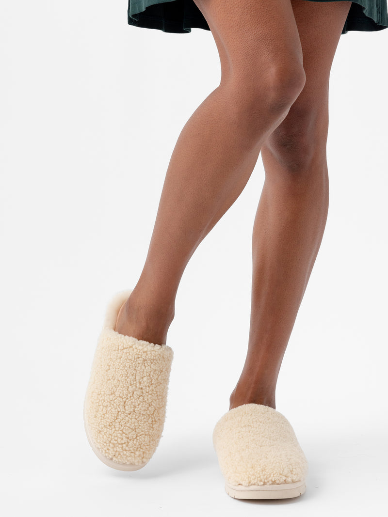 A person is wearing a dark green dress and standing with legs crossed at the ankles. Their feet are adorned in Cozy Earth's beige and fluffy Puffy Sheep Slippers, set against a white background.