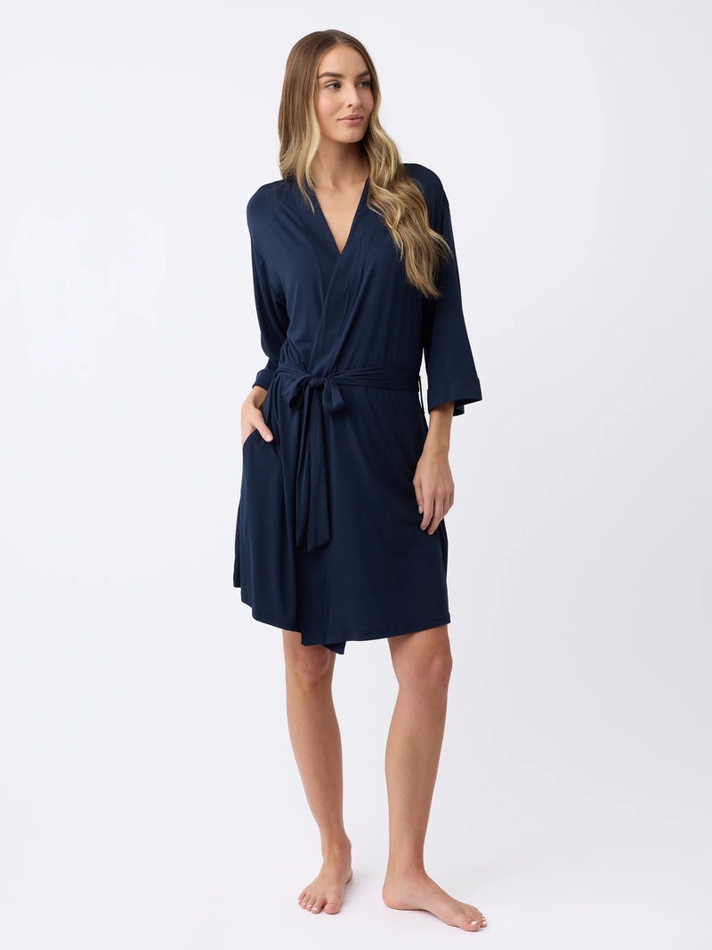 A person with long hair poses barefoot against a plain white background, wearing Cozy Earth's Women's Stretch-Knit Bamboo Kimono Robe in navy blue, featuring long sleeves, a wrap style, and a waist tie.