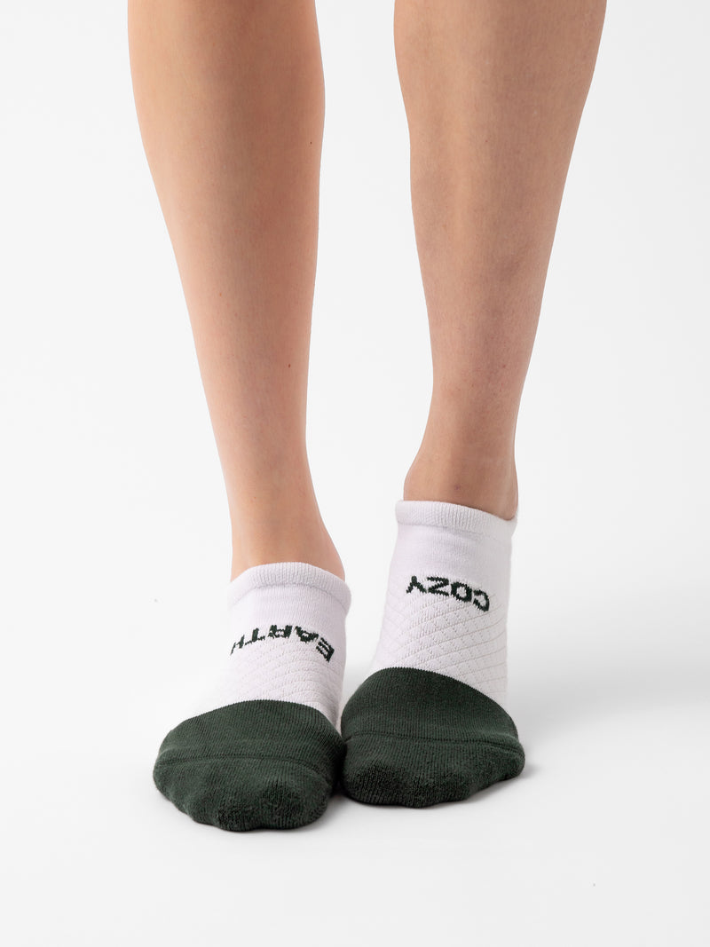 A person is wearing a pair of Cozy Earth's Essential Ankle Socks, featuring white uppers and dark green toes, standing against a plain white background. The socks display text near the ankle area, with "EARTH" on one sock and "COZY" on the other, although one word appears backward.