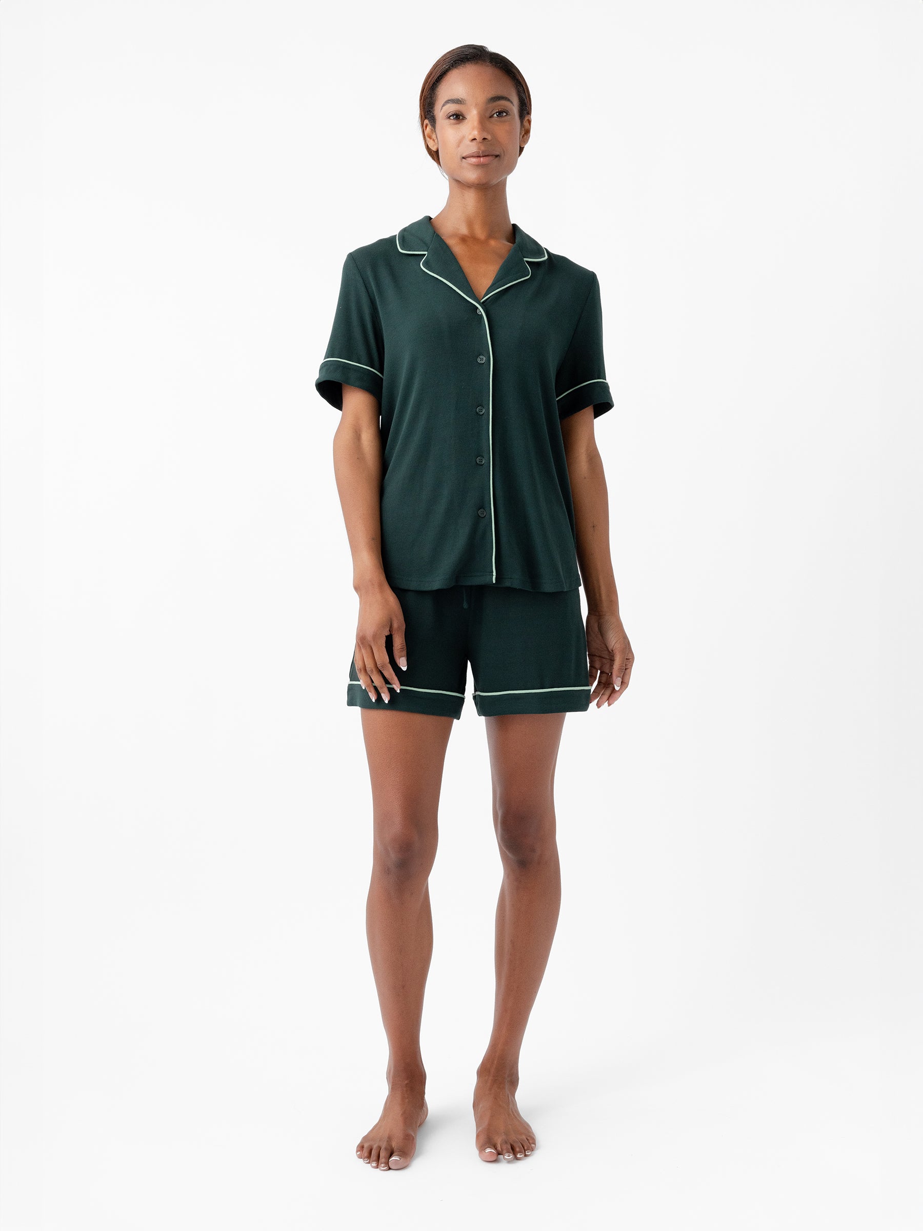 A person stands barefoot on a white background, wearing Cozy Earth's dark green Women's Bamboo Rib Knit Classic Pajama Short set, featuring a button-up short-sleeve top and matching shorts with light-colored piping along the edges. The person's arms are relaxed by their sides. 