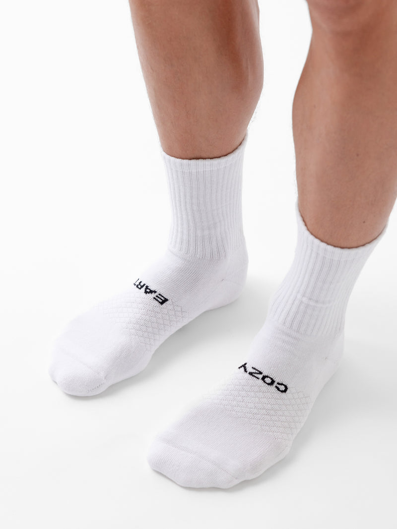 A person wears white Essential Calf Socks from Cozy Earth; the left sock reads "EARTH" and the right "COZY" in black letters. The background is plain white.