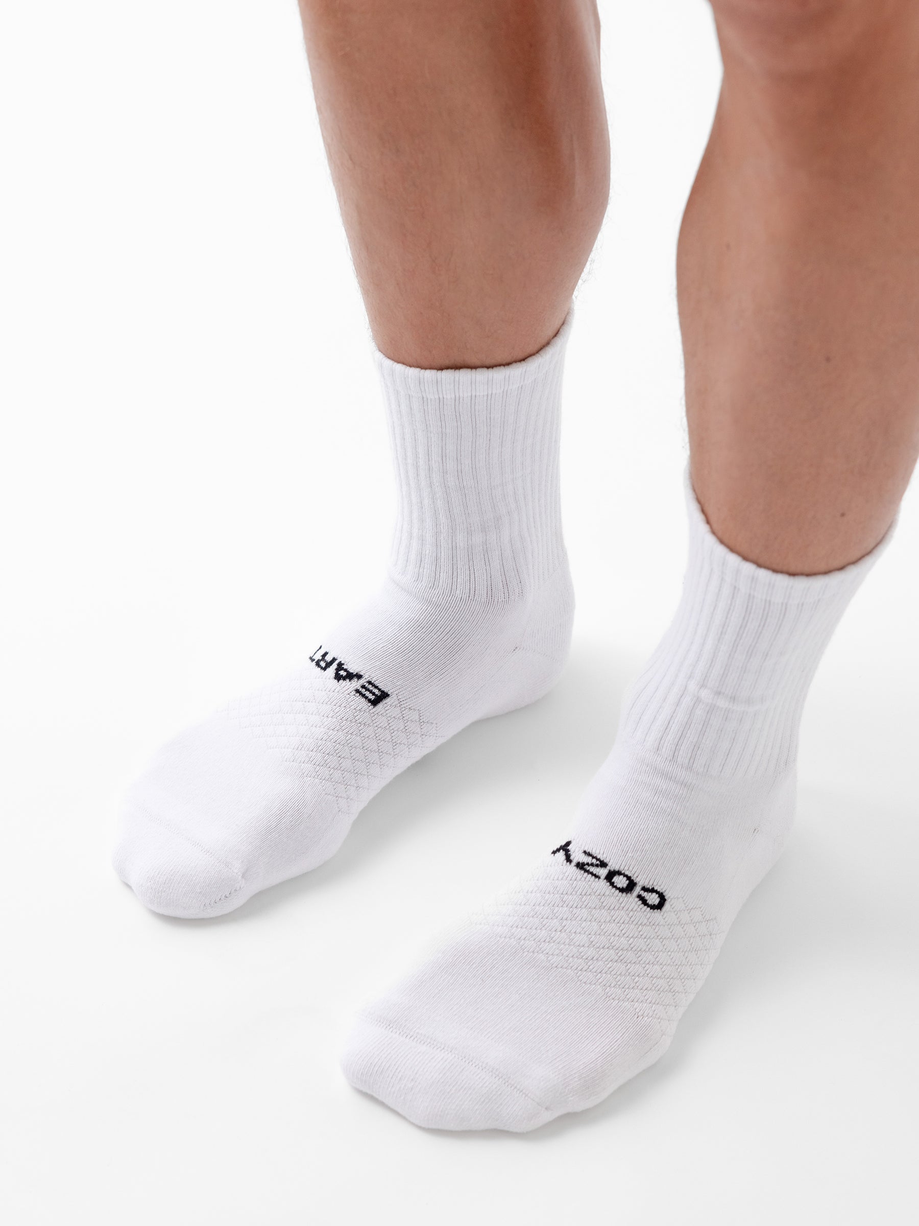 A person stands on a white surface wearing Cozy Earth’s Essential Calf Sock 8-Pack in white. The left sock reads "EARTH" and the right reads "COZY" in black letters near the toes. 