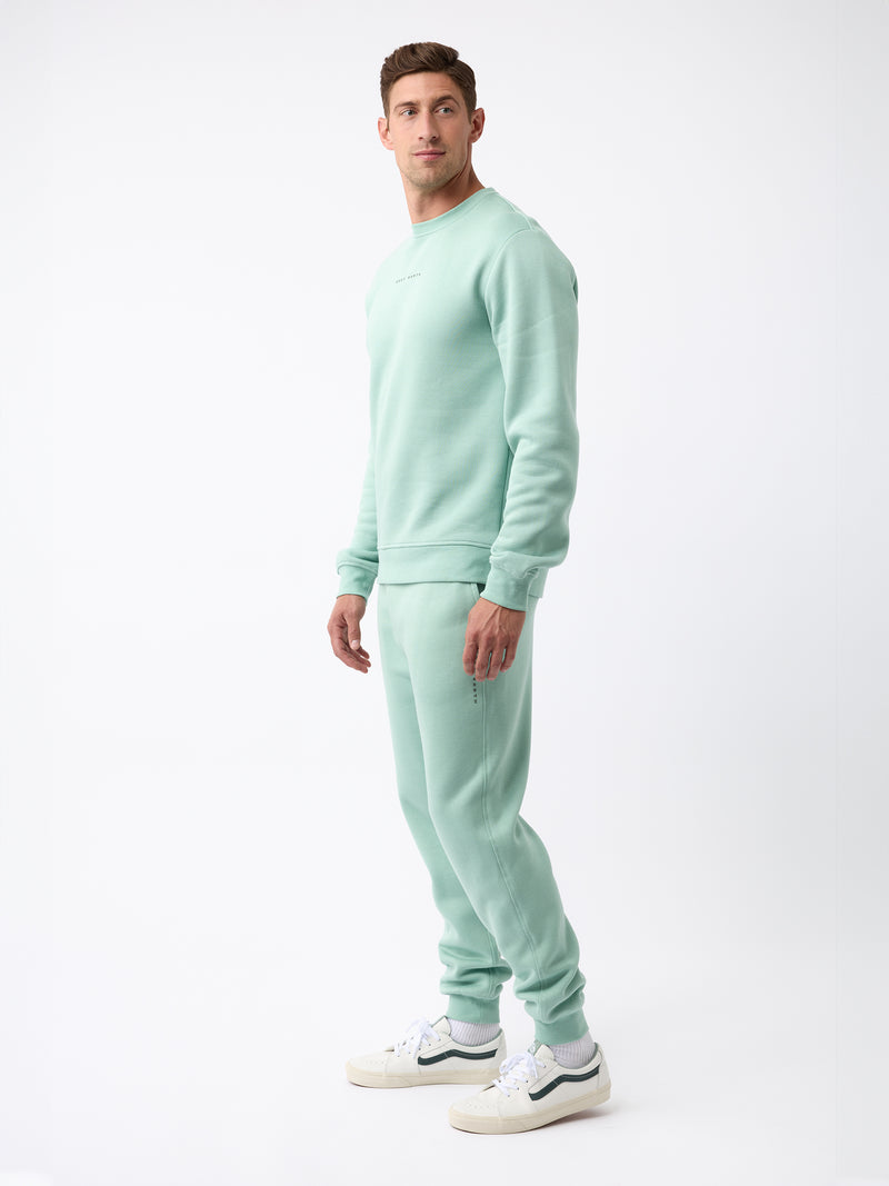 A person stands against a white background, dressed in the Men's CityScape Jogger from Cozy Earth, along with white sneakers. The individual looks at the camera with a neutral expression, and the jogger features minimal branding.