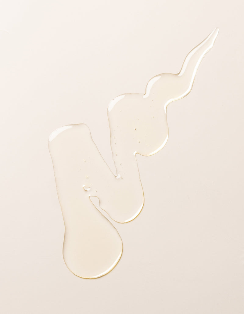 A swirl of transparent, light-colored Cozy Earth's Revitalizing Pair liquid on a plain, pale beige surface. The liquid forms irregular shapes with droplets at the ends, creating a visually interesting pattern.