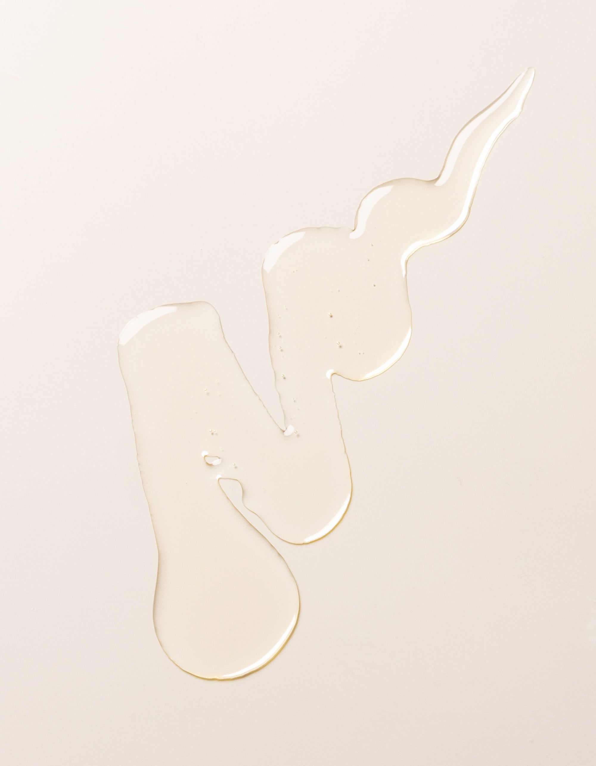 An irregular-shaped spill of Cozy Earth's Oil Cleanser on a smooth, light-colored surface. The clear liquid forms a long, winding pattern with droplets and uneven edges, creating the impression of fluid motion.