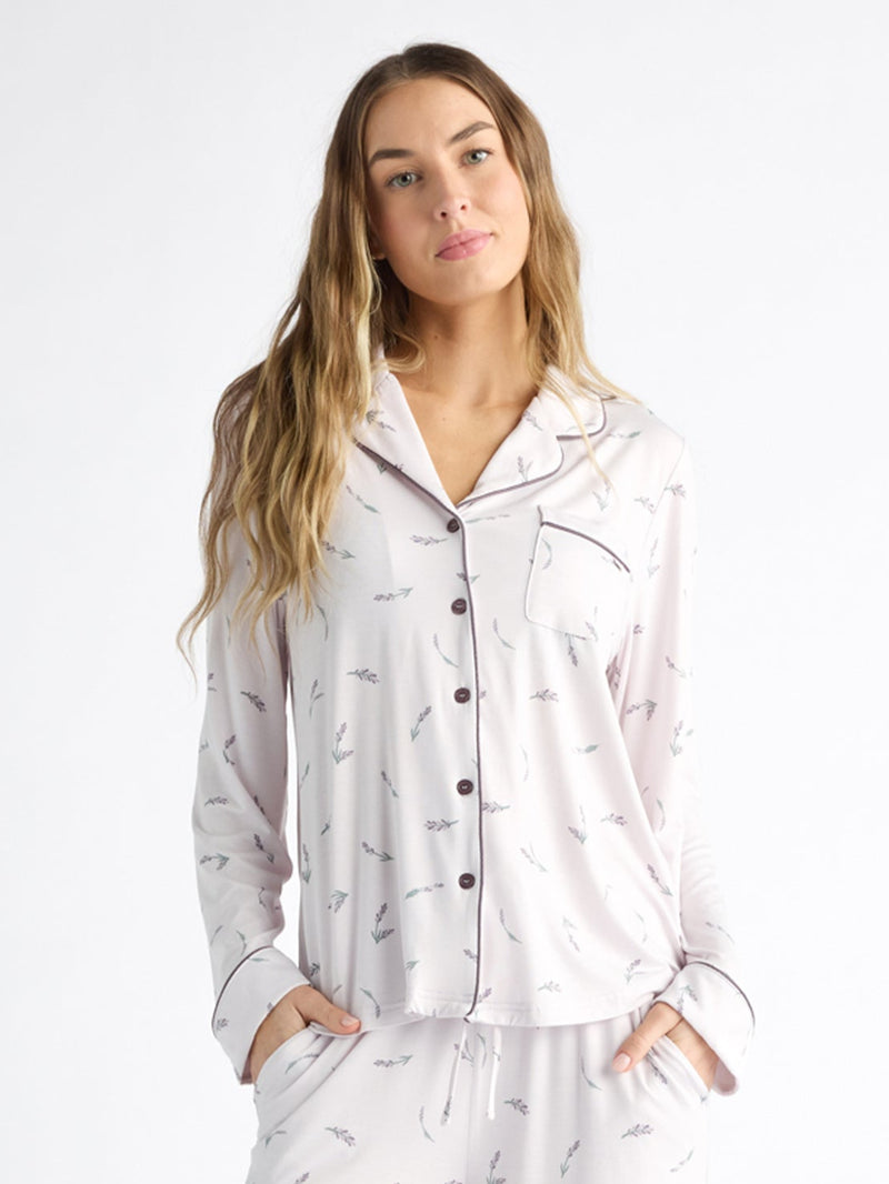 A person wearing Cozy Earth Women's Bamboo Stretch-Knit Long Sleeve Pajama Top, featuring a collar, buttons, and chest pocket, stands relaxed against a white background with long hair and hands in pockets. The light-colored top has a subtle print.