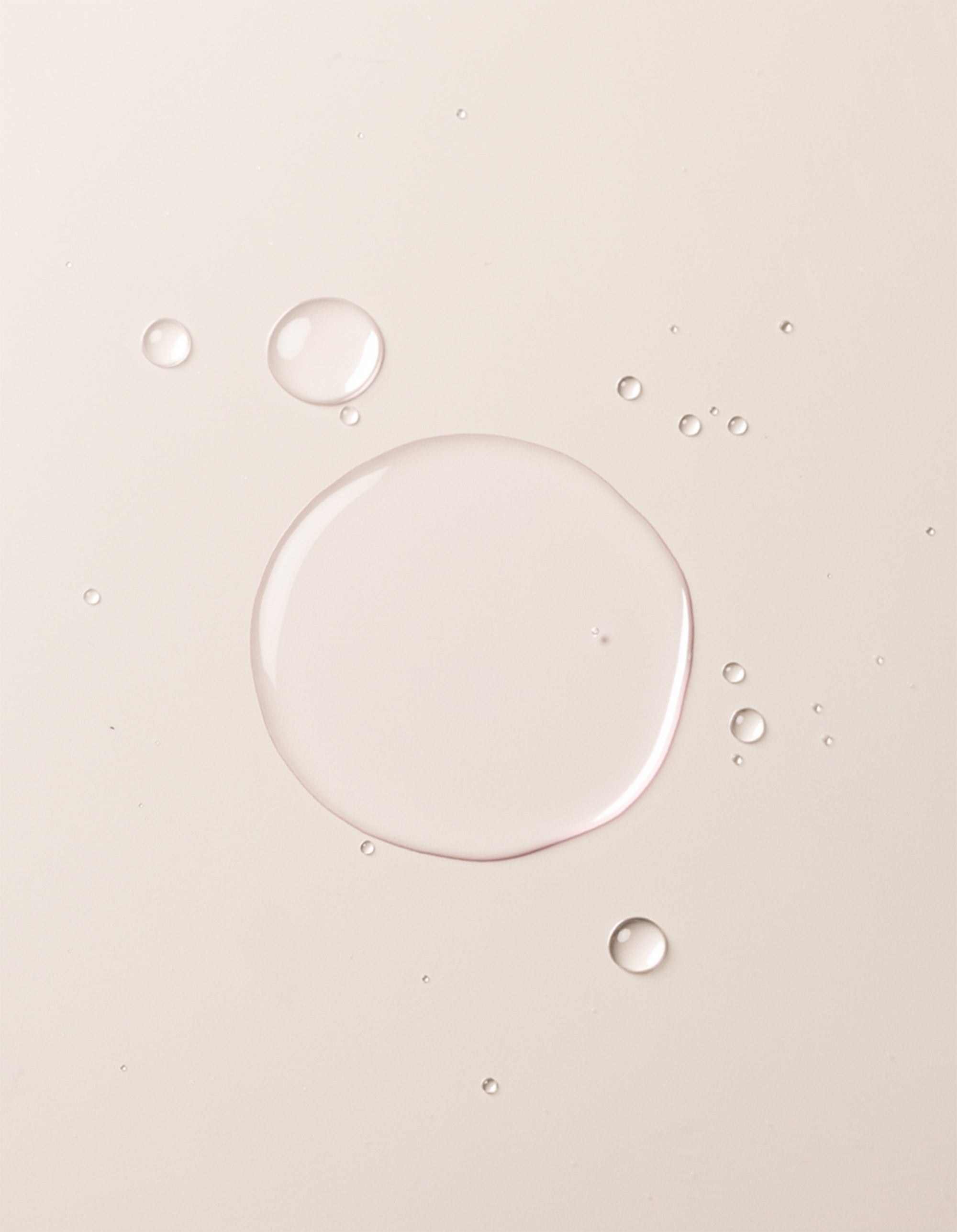 A close-up image showcases translucent liquid droplets of Cozy Earth's Balancing Face Mist on a smooth, off-white surface. The droplets vary in size, with one large central droplet surrounded by several smaller ones, creating a clean and minimalistic appearance. 