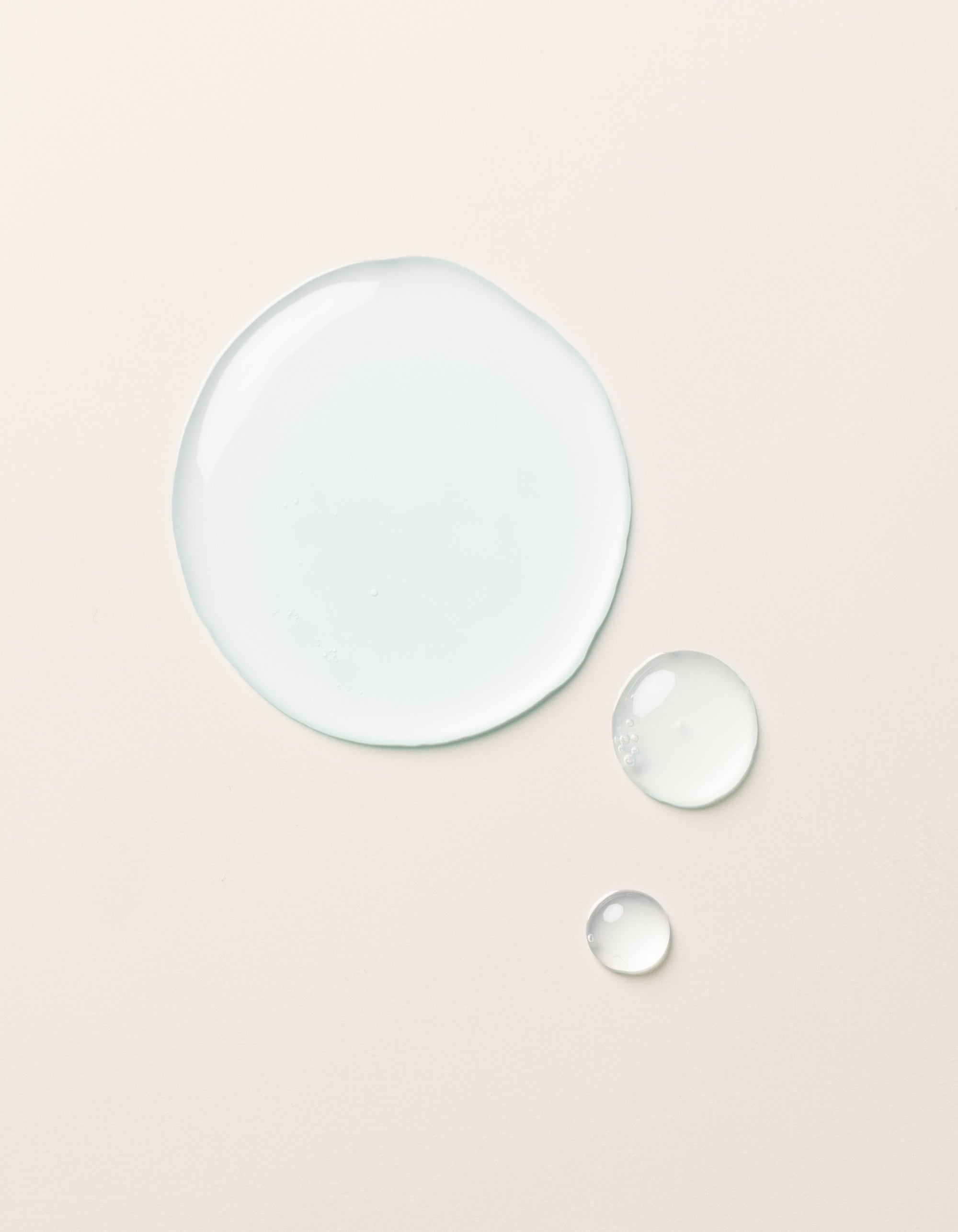 A large clear droplet and two smaller droplets of Cozy Earth Hydrating Serum rest on a smooth, light beige surface. The glossy, semi-transparent droplets reflect light slightly on their surfaces.