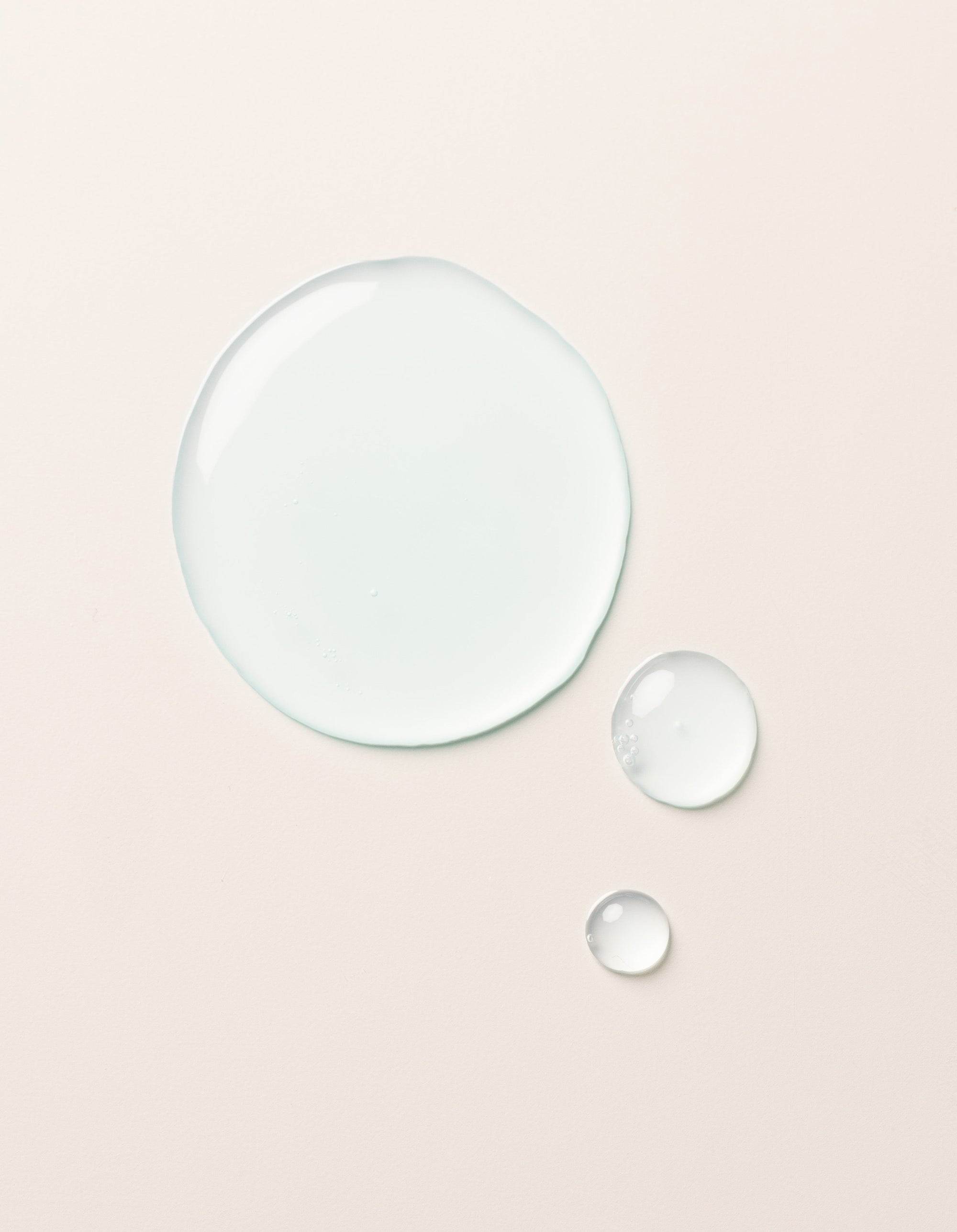 The Hydration Duo by Cozy Earth is shown as a large drop and two smaller drops of clear, light blue liquid on a smooth, light beige surface. The drops vary in size but all maintain a rounded shape, appearing glossy and slightly reflective.