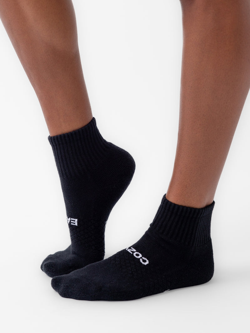 A person wearing black, quarter-length socks stands against a white background. The socks, from the Essential Quarter Sock 2-Pack by Cozy Earth, feature a ribbed top and textured pattern with the brand name "COZY" visible on the side. The person's skin is light brown.