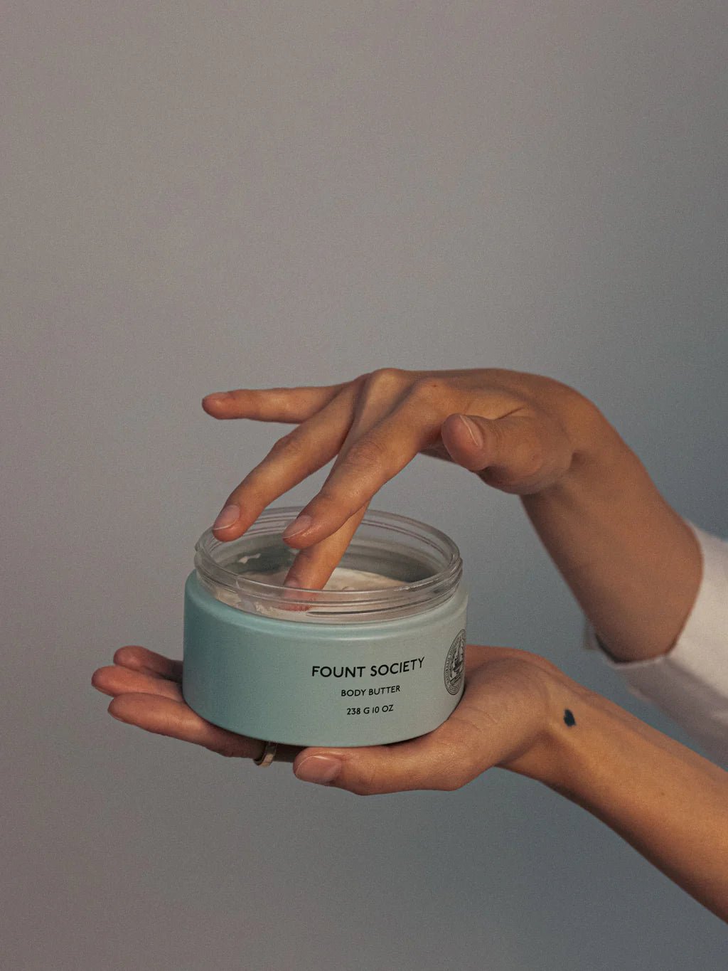 A woman holds Fount Society Body Butter in her hand and dips one finger into the lotion.