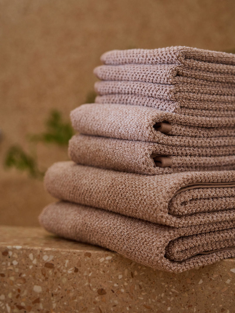 A stack of Nantucket towels and washcloths in Heathered Clay sit on the edge of a stone bathtub.
