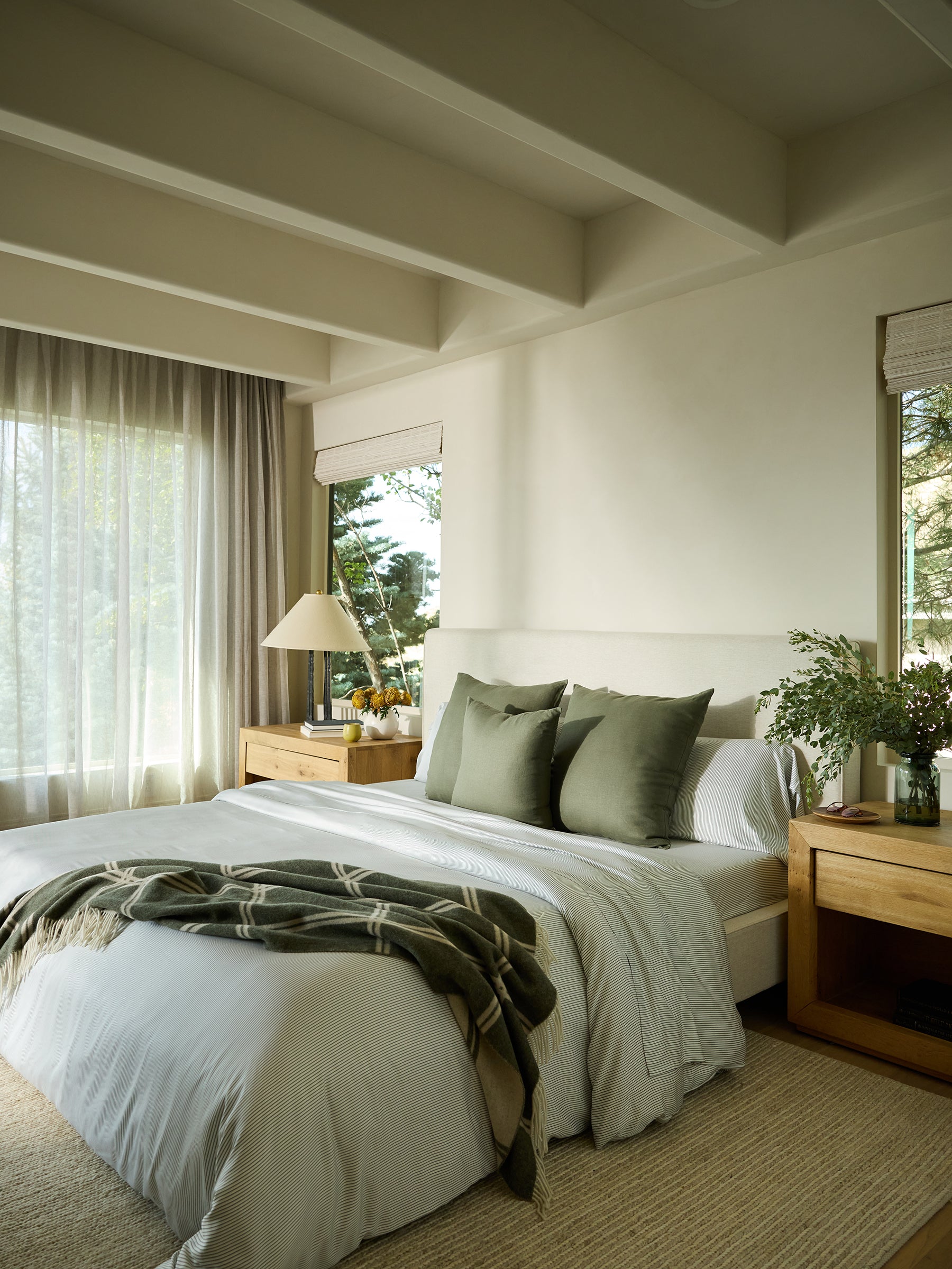 A tranquil bedroom features a spacious bed draped in Cozy Earth's Bamboo Sheet Set, showcasing a crisp white and green design. Two matching green pillows and a checkered blanket add charm to the bed. Wooden nightstands with lamps and vases grace each side, while sunlight pours through sheer curtains framing the window. 