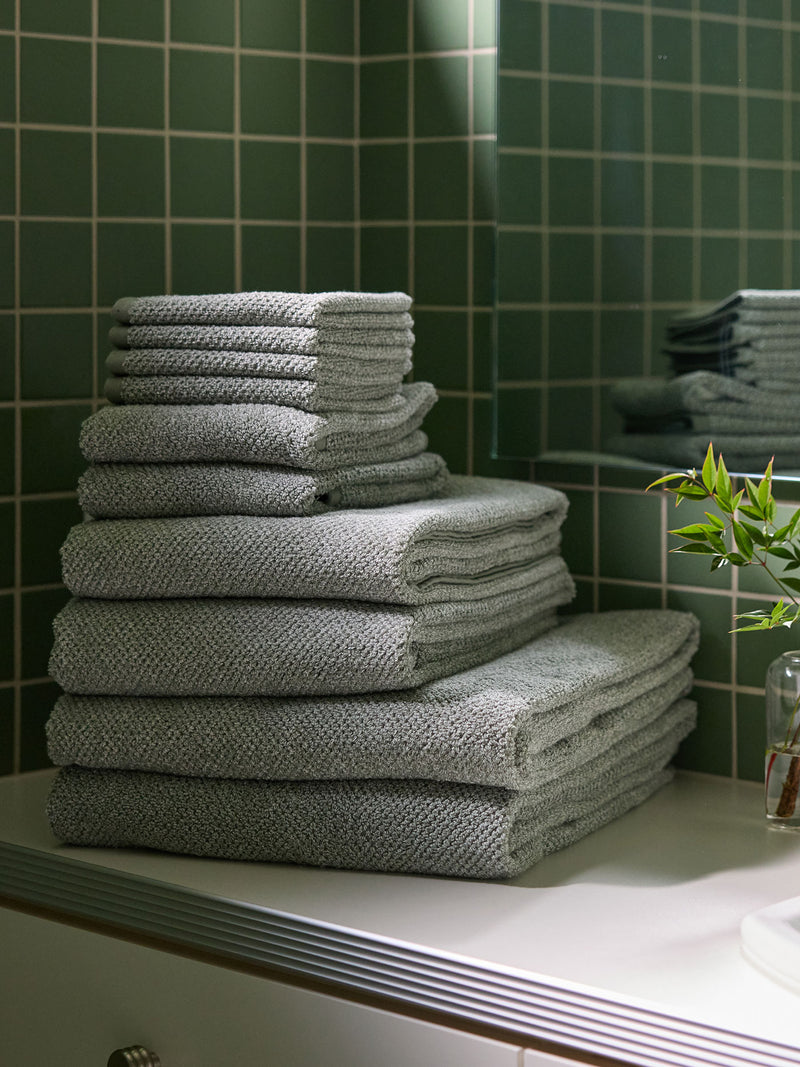 A stack of Nantucket towels in various sizes sit on the counter of a bathroom vanity. The wall behind is tiled in green tile.