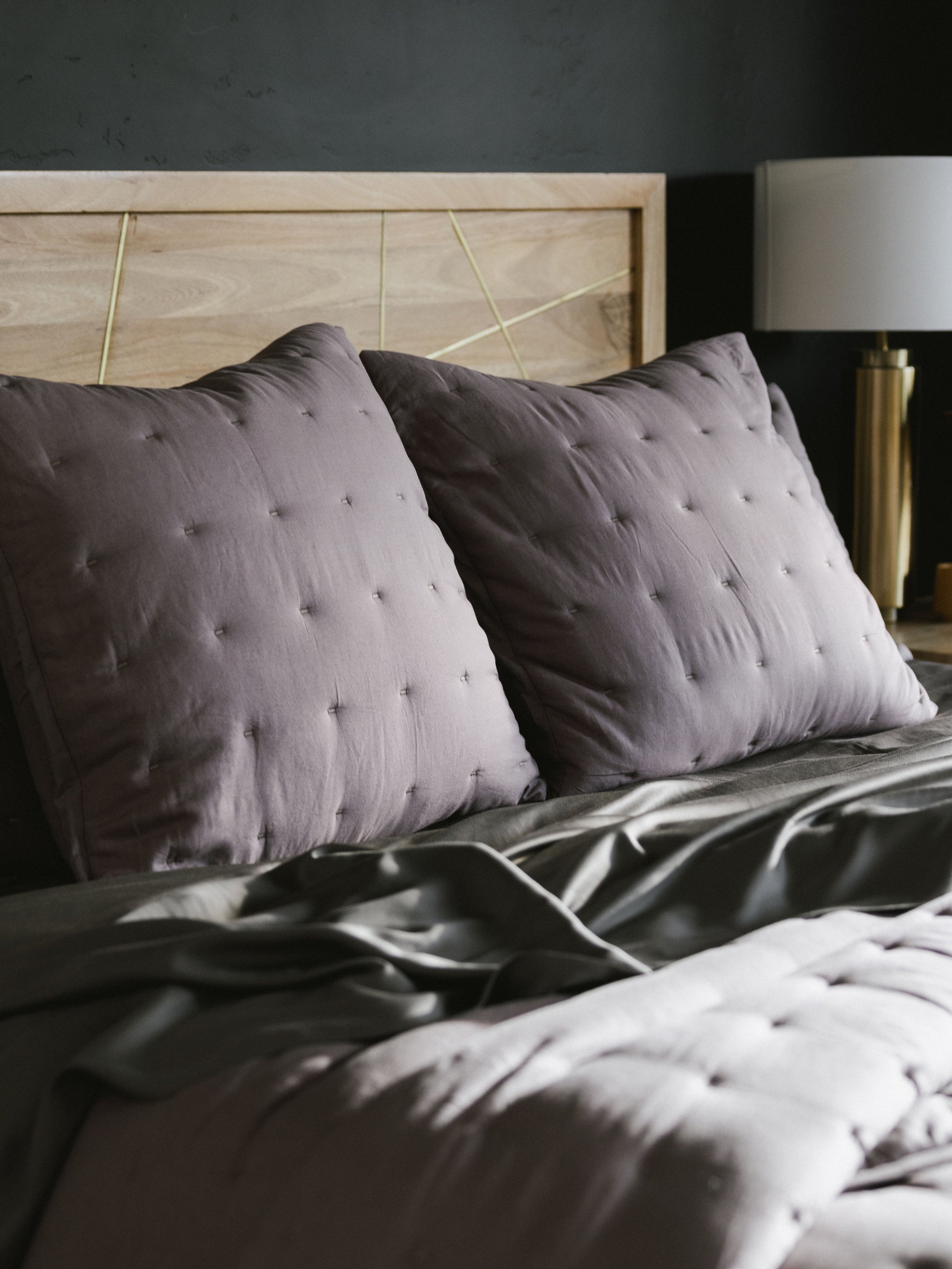 A cozy bed with Cozy Earth's Premium Down Alternative Insert covered in dark gray bedding and adorned with two large, tufted gray pillows rests against a light wood headboard. On the right side, a modern bedside lamp with a gold base adds elegance to the scene. The room's dark wall background creates a striking contrast with the light-colored headboard and pillows. 