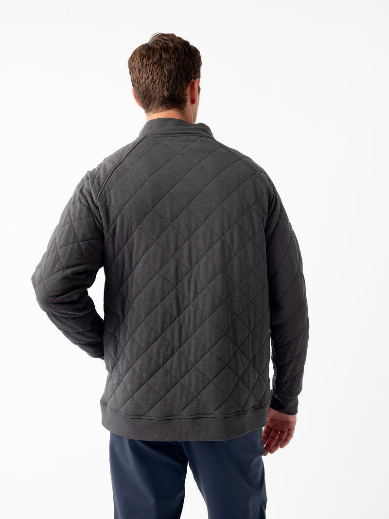 A man with brown hair is standing with his back to the camera, wearing a gray Men's Ultra-Soft Bamboo Quilted Snap Pullover from Cozy Earth and dark blue pants. His left hand is positioned on his hip. The background is plain white.