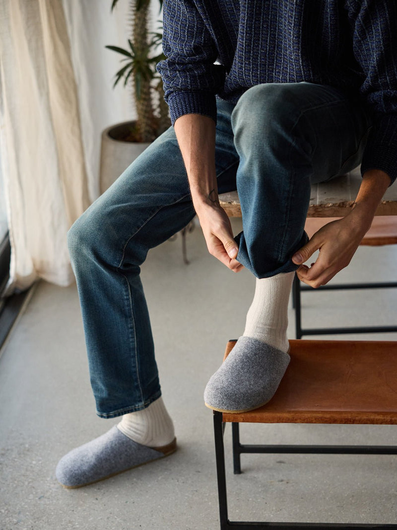 A person in a blue sweater and jeans sits on a chair, adjusting their pant leg. They're wearing white socks with gray Lakehouse Clogs by Cozy Earth. A plant and curtain are visible in the background.