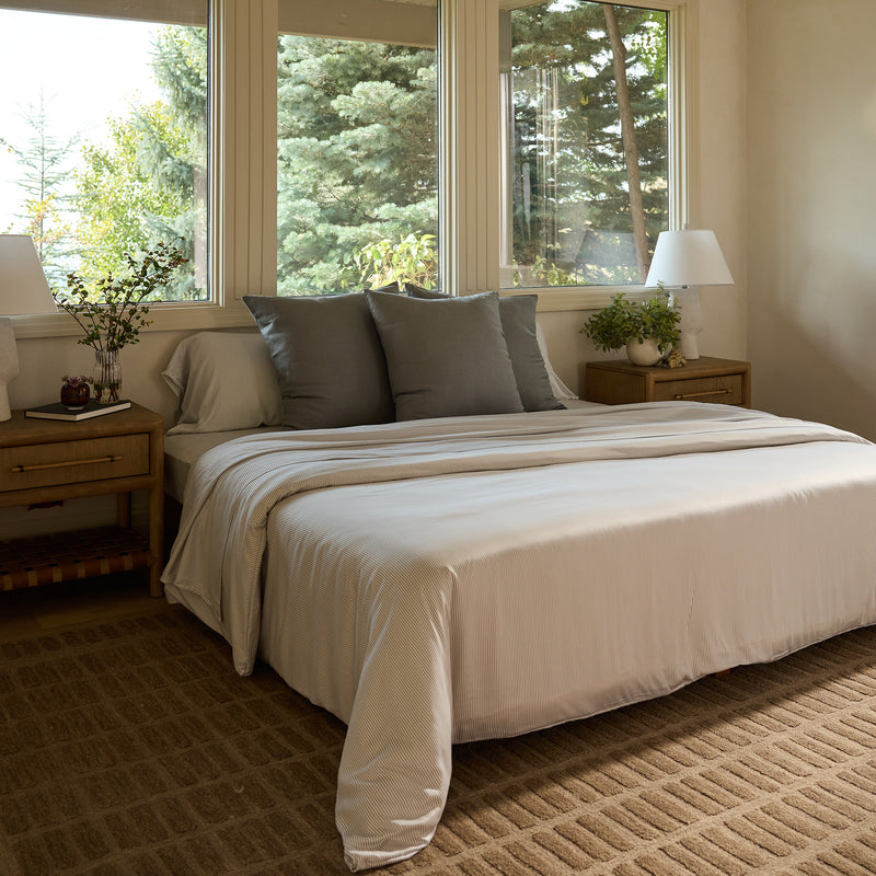 A cozy bedroom showcases a large bed adorned with Cozy Earth's Bamboo Sheet Set in white and gray. On either side of the bed, two wooden nightstands hold white lamps. Large windows offer a view of lush green trees, while a brown rug adds warmth to the floor.