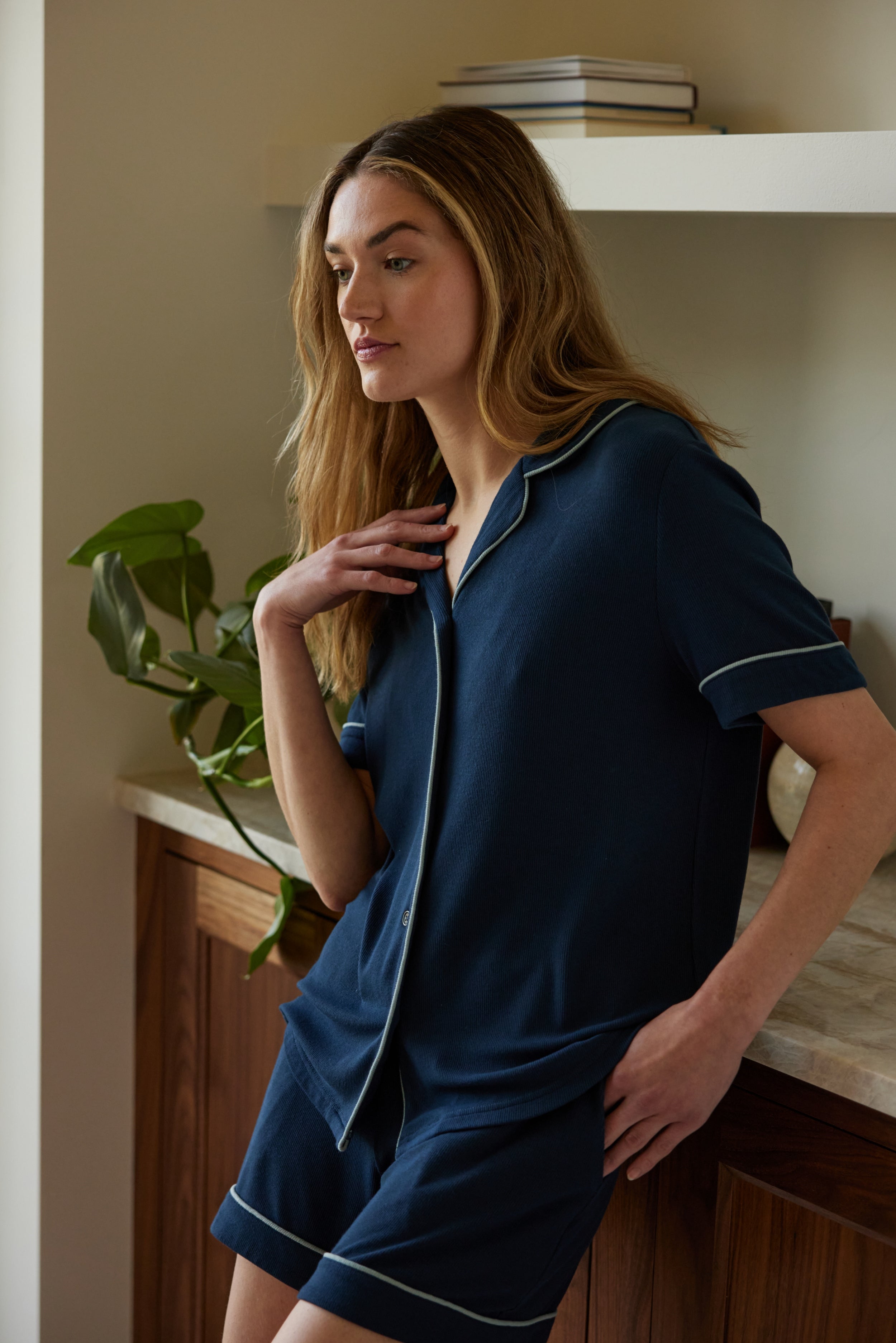 A woman with long hair leans against a wooden counter, wearing the Cozy Earth Women's Bamboo Rib Knit Classic Short Sleeve Pajama Top in navy blue with white piping. She gazes thoughtfully to the side, resting one hand on her chest and the other in her pocket. A potted plant and books are visible in the background. |Color:Eclipse