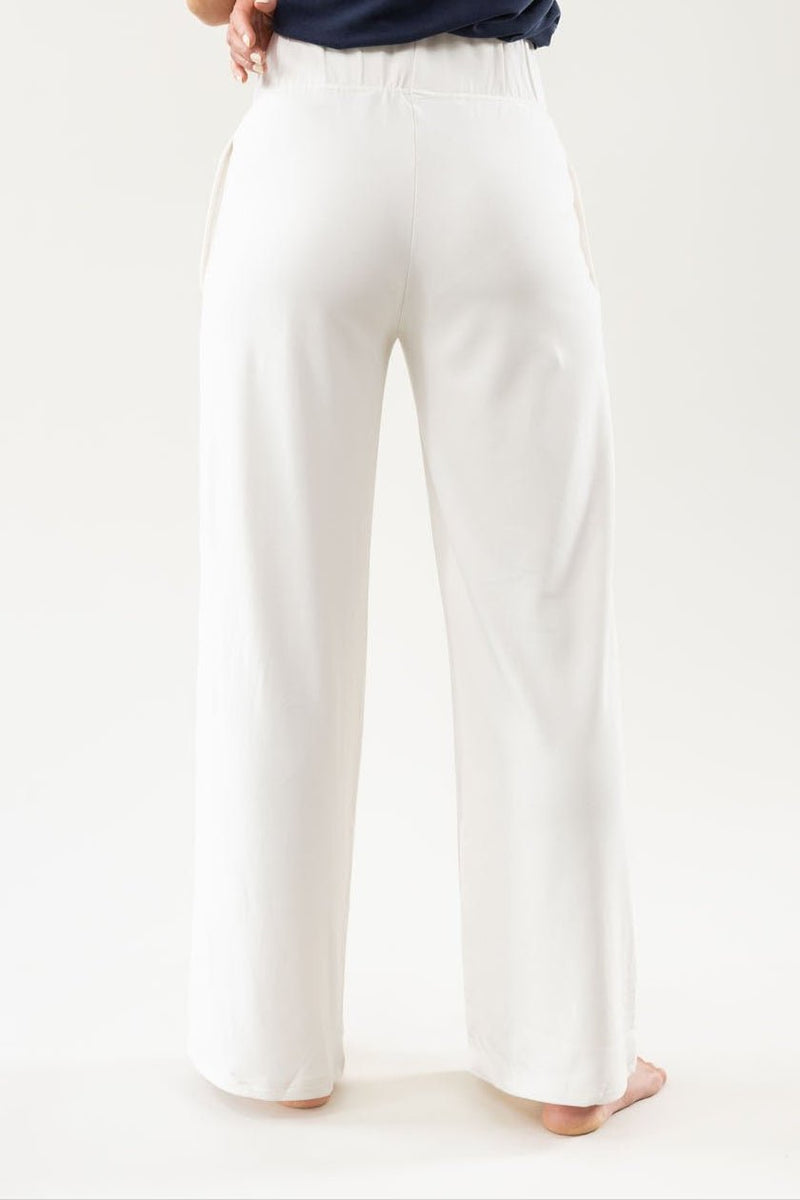 A person stands barefoot with their back to the camera, wearing a navy top and loose-fitting white pants from the Women's Ultra-Soft Bamboo Wide Leg Pull On Pant & Pullover Set by Cozy Earth. The background is plain and light-colored.