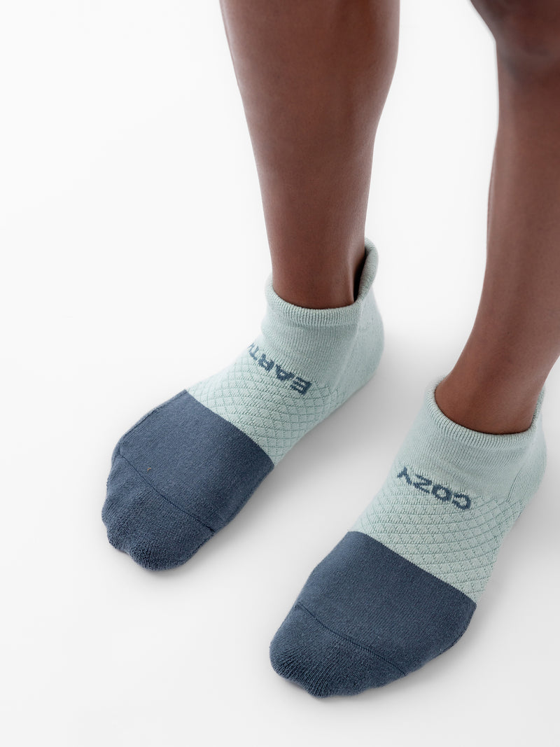 Close-up of a person wearing green and dark gray Cozy Earth Essential Ankle Socks standing on a white surface. The socks have text, "COZY" and "EARTH" woven into the fabric on the tops of the feet.
