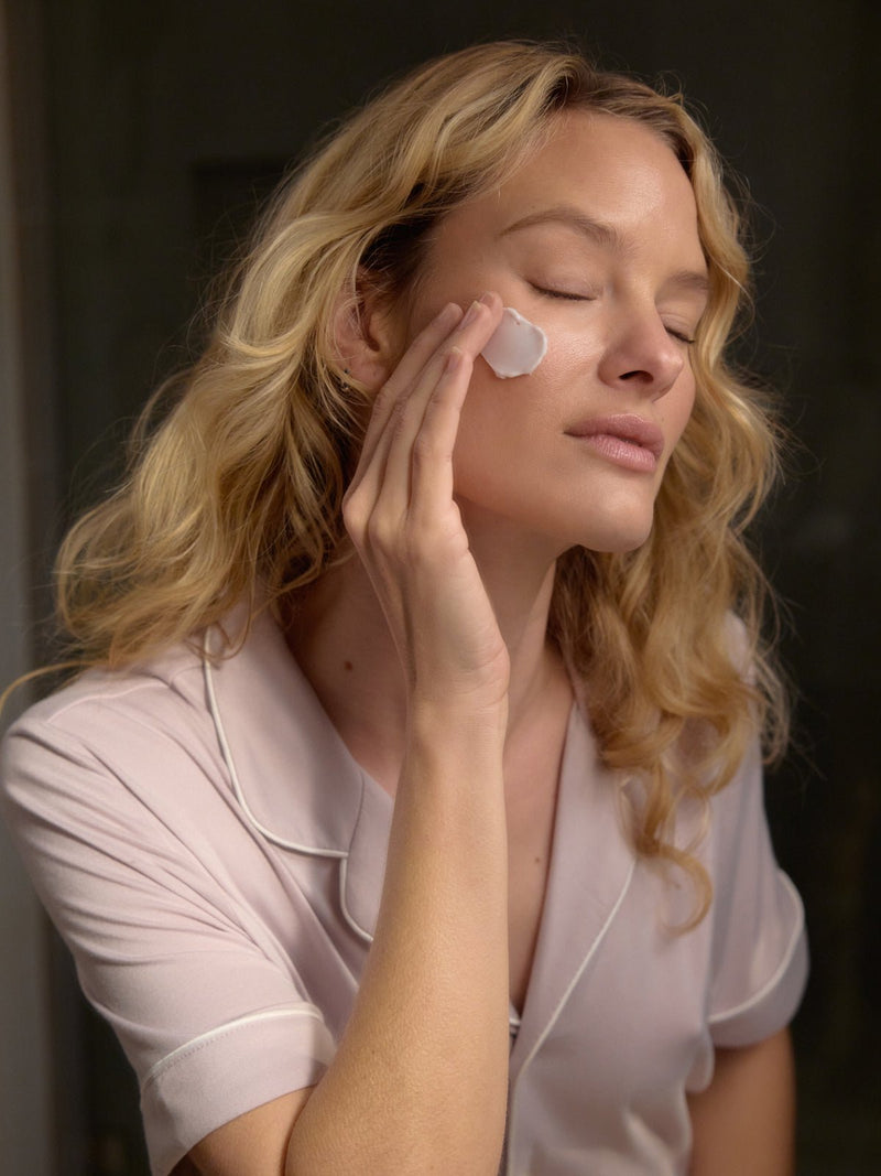 A person with long blonde hair applies a dab of cream to their cheek from the Rest & Reset Bundle by Cozy Earth. Wearing a pastel pink pajama top, their eyes are closed in relaxation. The dimly lit background highlights their calm expression and skincare routine.
