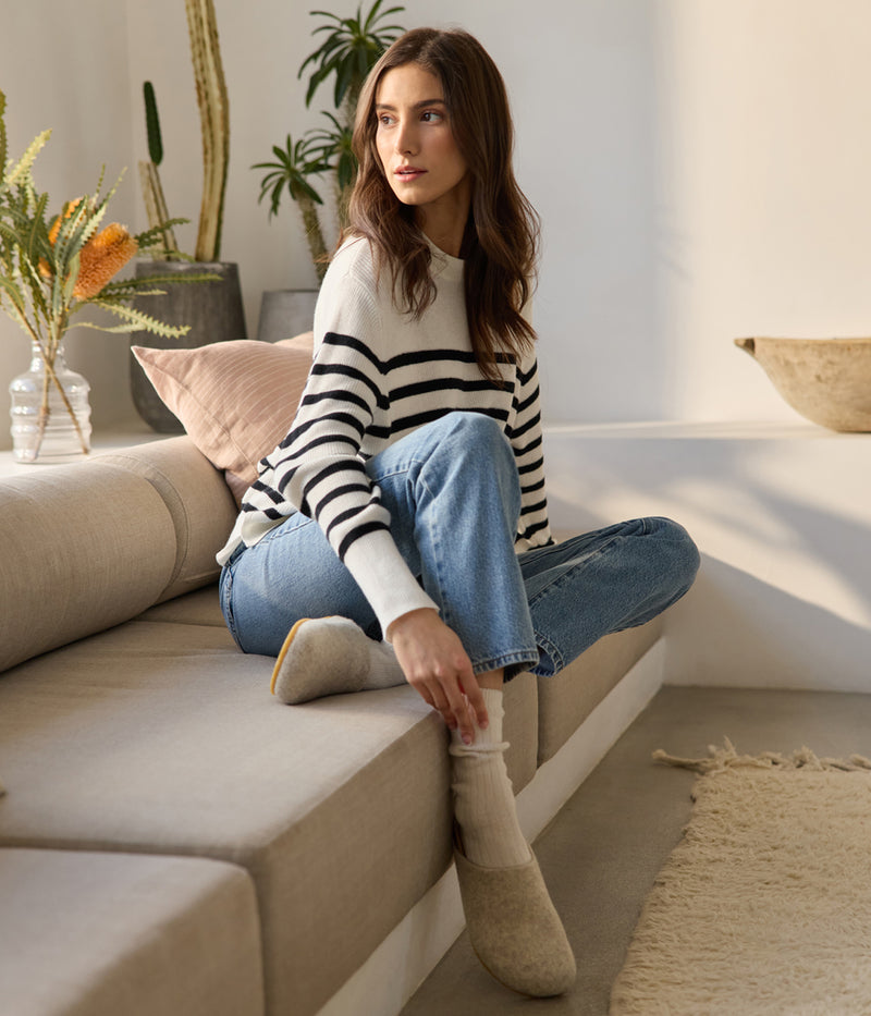 A woman lounges on a beige sofa, sporting a white and black striped sweater, blue jeans, and Lakehouse Clog boots by Cozy Earth. She gazes sideways as potted plants and a textured rug enhance the room's cozy ambiance.