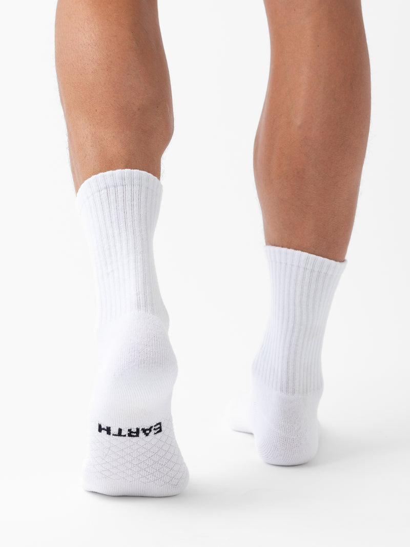 A person stands on tiptoes, viewed from behind, wearing white ribbed Essential Calf Socks by Cozy Earth. “EARTH” is printed in black on the sole of the left sock. The background is plain white.