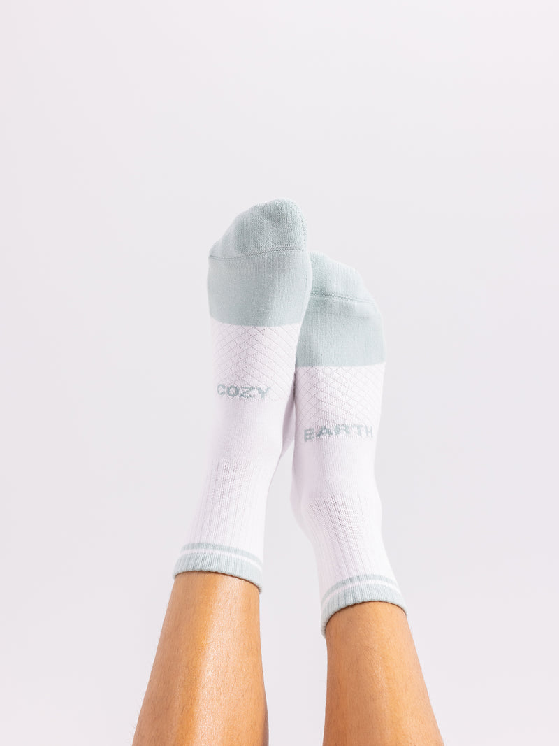 Two feet are clad in socks from the Essential Quarter Sock 4-Pack by Cozy Earth. The socks are white with pale green toes, heels, and tops, featuring "COZY" on one and "EARTH" on the other. The legs are lifted against a plain white background.